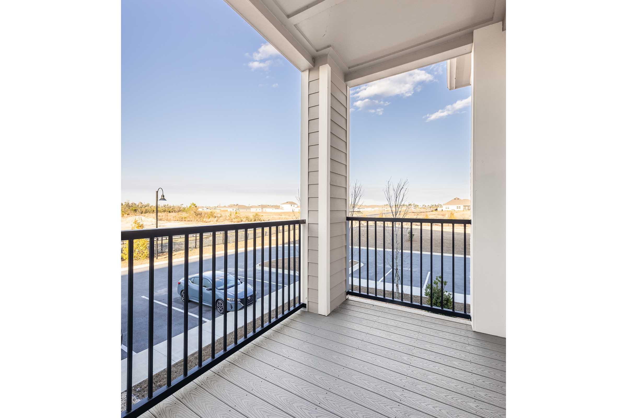 A spacious balcony with a wooden floor and black metal railing. The view features a clear sky and distant landscape, with a parking lot visible below. A single car is parked nearby, and there are some sparse trees and vegetation in the background.