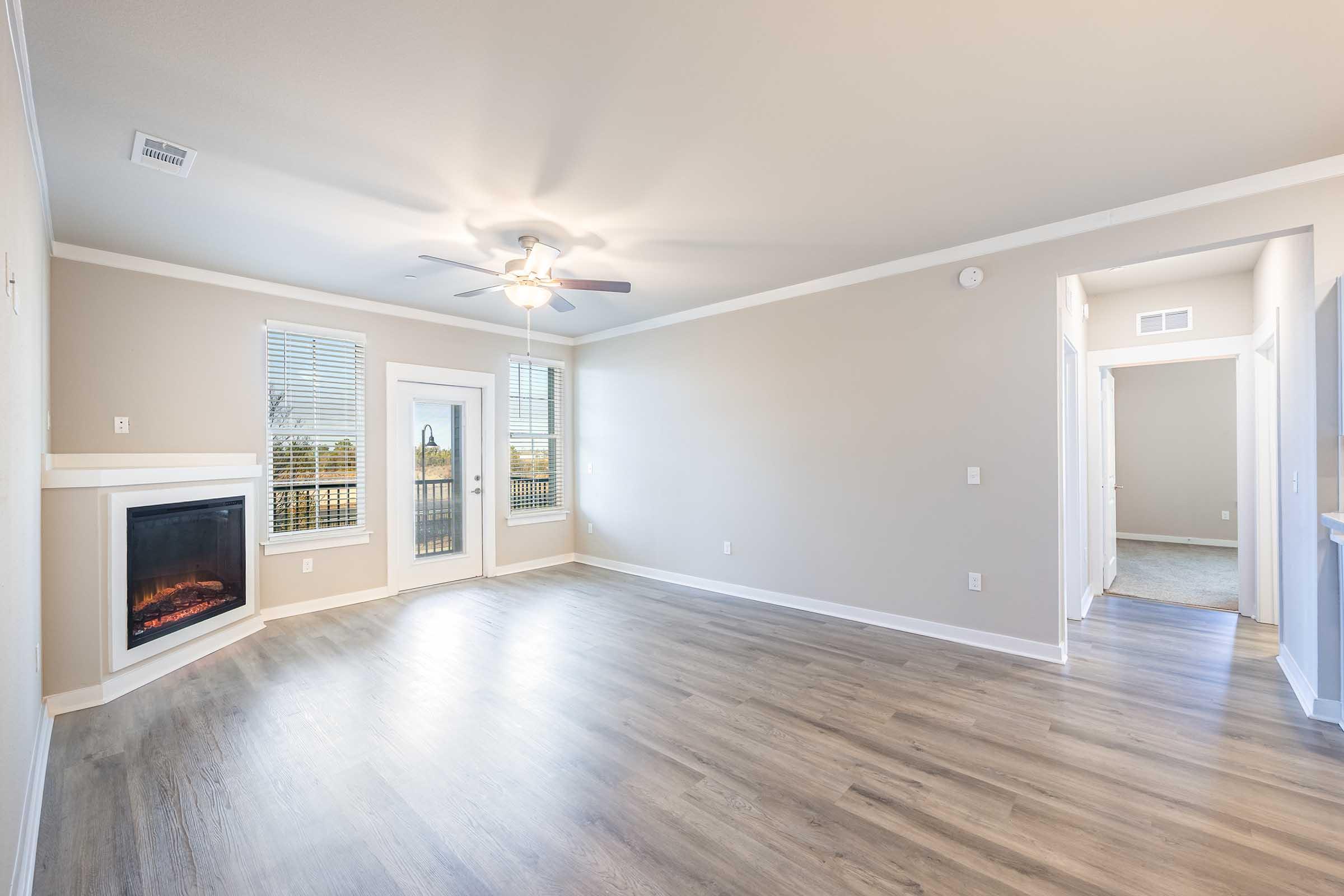 Bright and spacious living room with light-colored walls and wooden flooring. Features a ceiling fan and an electric fireplace. Large windows provide natural light, and a door leads to a balcony. A hallway is visible on the right, leading to other rooms.