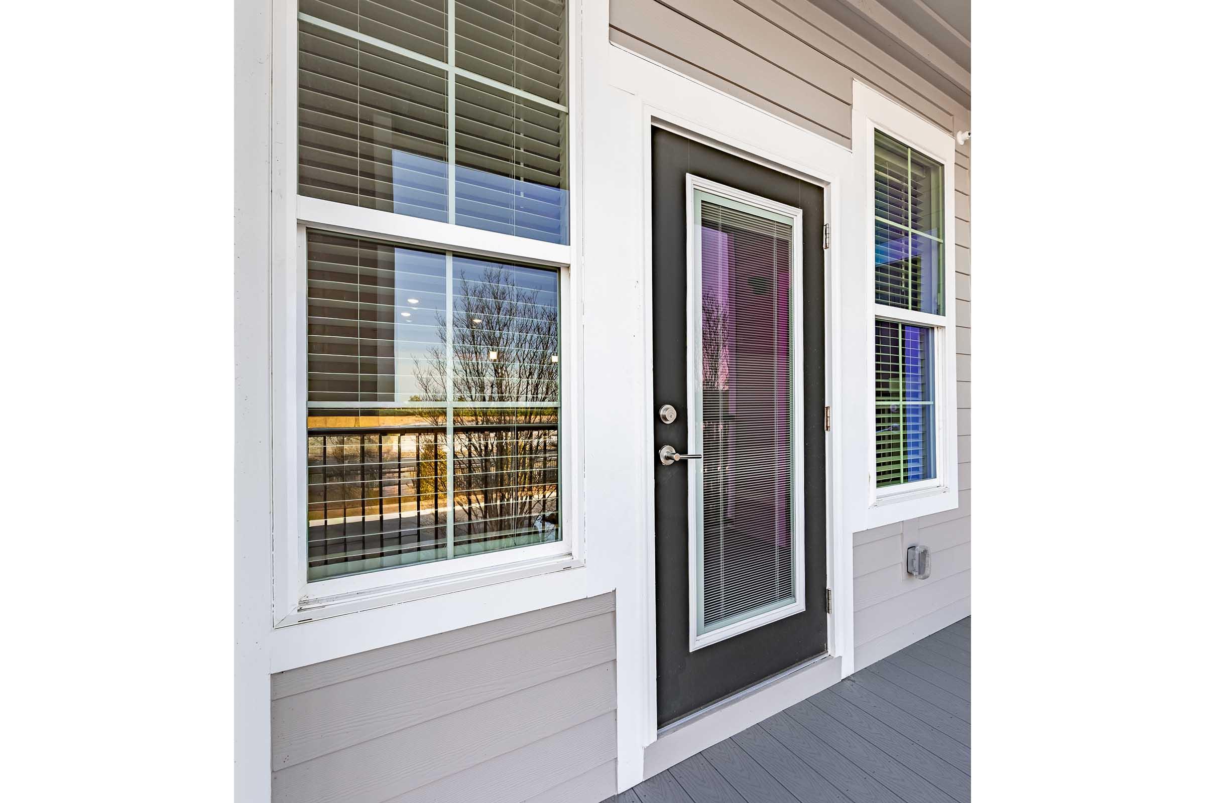A modern front door with a glass panel and vertical blinds, surrounded by large windows. The door is dark in color, while the house exterior features light-colored siding. Natural light is visible through the windows, and there are decorative railings on a small porch area.