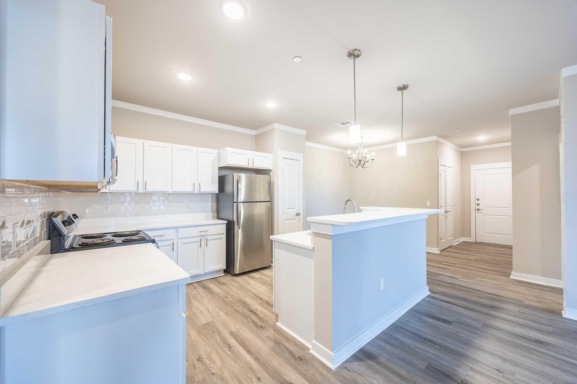 A modern kitchen featuring white cabinets, stainless steel appliances, and a large island with a sink. The space is brightly lit with overhead lights and has an open layout leading to a dining area. Light-colored walls and wood-like flooring add to the contemporary aesthetic. 