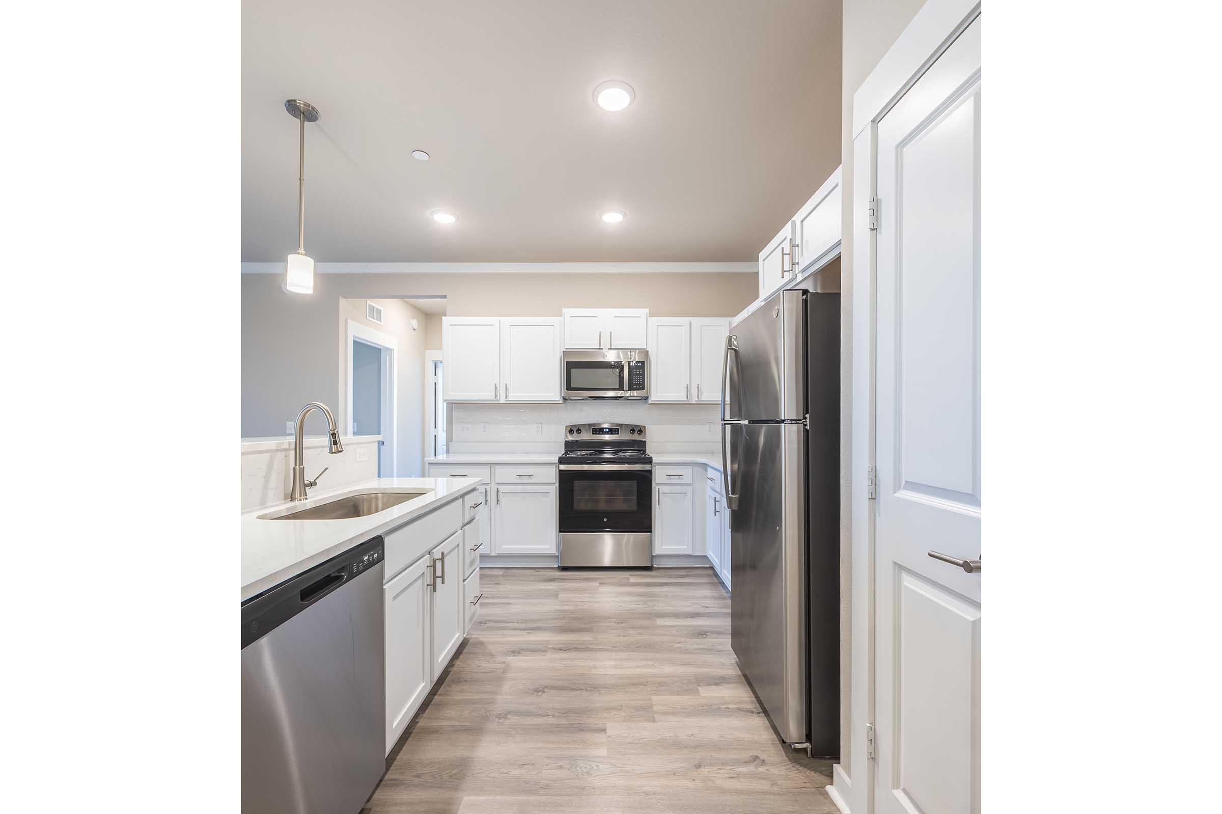 Modern kitchen featuring white cabinetry, stainless steel appliances including a fridge, stove, and dishwasher. The space has a light-colored countertop, hardwood flooring, and recessed lighting, creating a bright and inviting atmosphere. The layout is open, showcasing a clean and functional design.