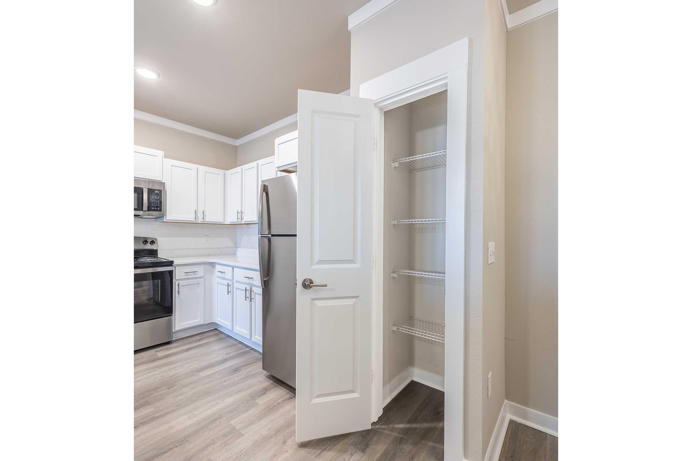 A modern kitchen featuring white cabinetry, stainless steel appliances, and a pantry closet with wire shelving. The pantry door is open, revealing the organized shelves inside. The flooring is light-colored, providing a warm and inviting atmosphere. The walls are a soft beige, complementing the overall design.