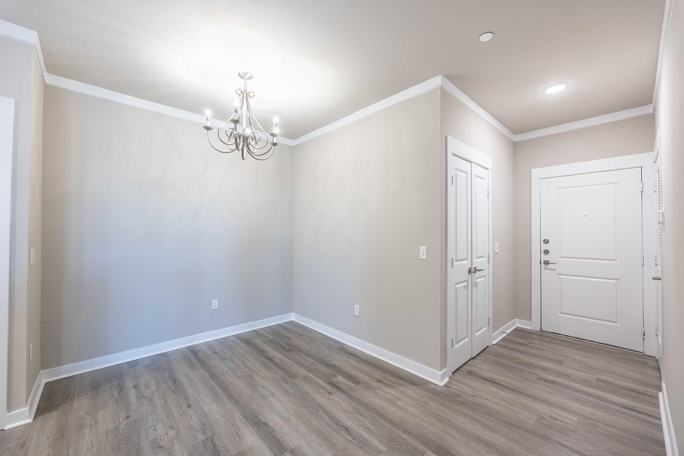 A well-lit, modern entryway featuring light gray walls, hardwood-like flooring, and a decorative chandelier. The entrance door is white, with a small window beside it. There is a closet door on the left, creating a minimalist and welcoming atmosphere.