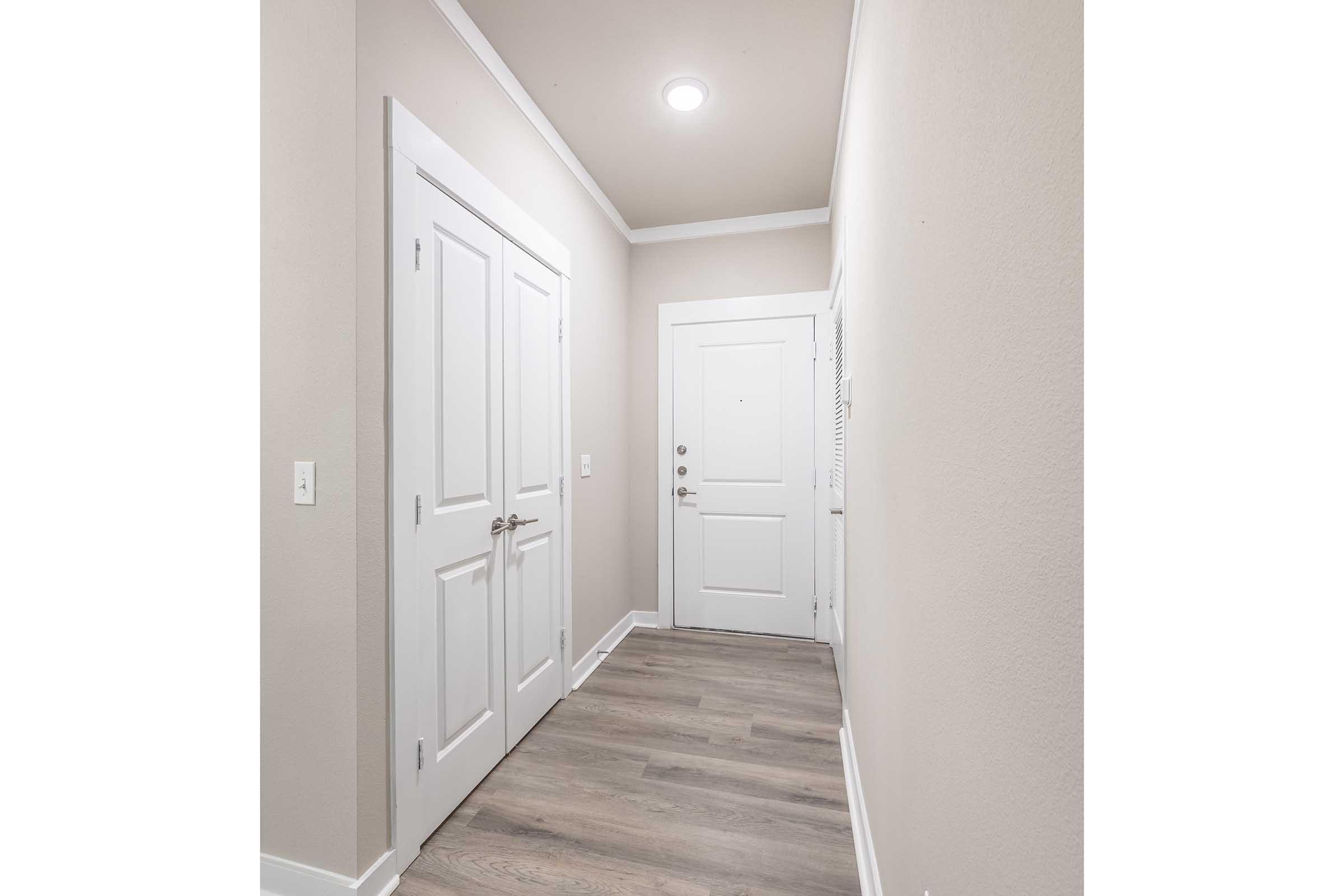 A well-lit hallway featuring light gray walls, a white ceiling, and a wooden floor. The space includes a closed white door on the right, a double-door closet on the left, and a door leading outside at the end of the hallway. The overall design is modern and clean.
