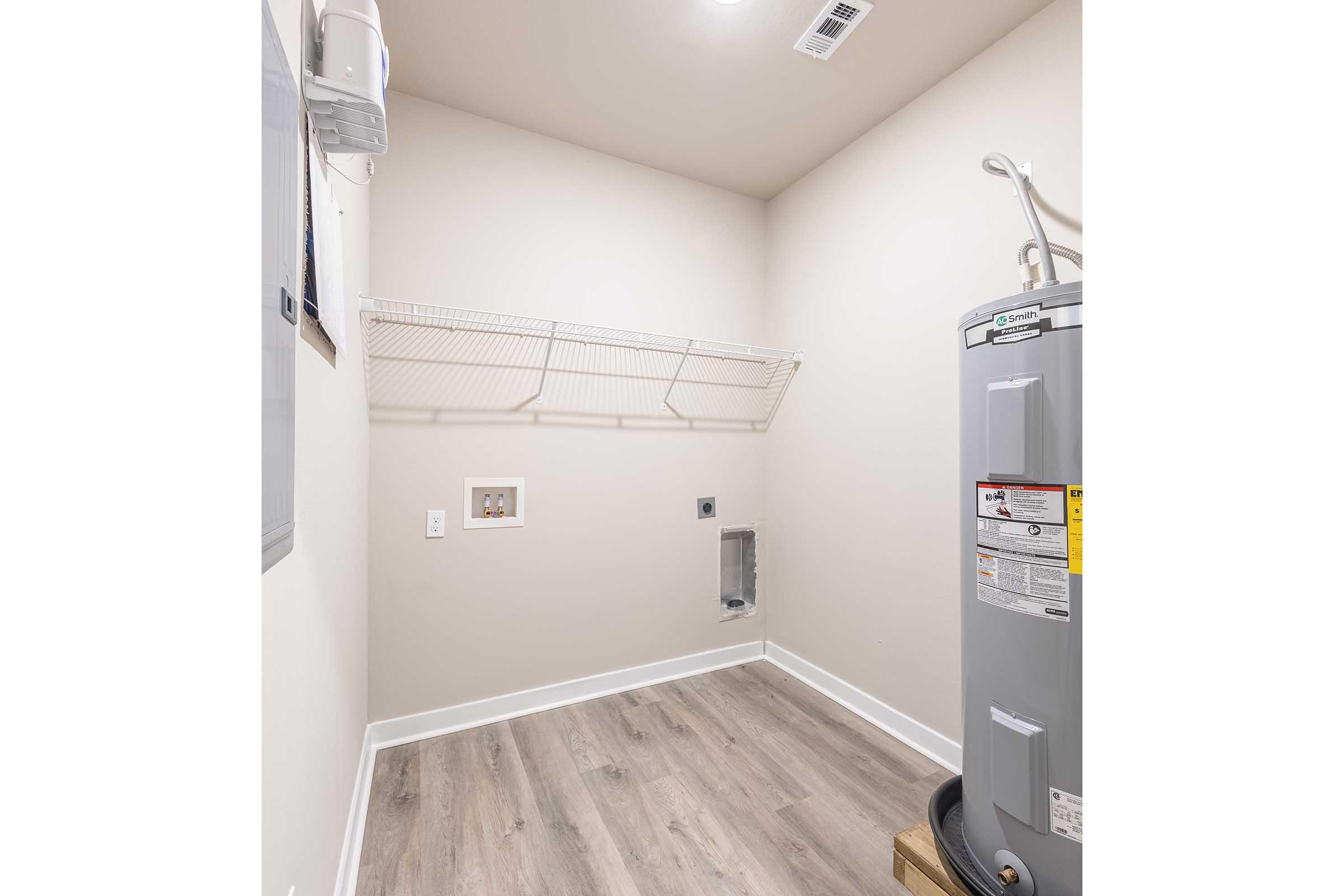 A clean, minimalist laundry room with light-colored walls and a light wood floor. Features a gray water heater on the right and a wire shelving unit on the left, with electrical outlets visible. There is a space for appliances, but no machines are present. The room is well-lit and uncluttered.