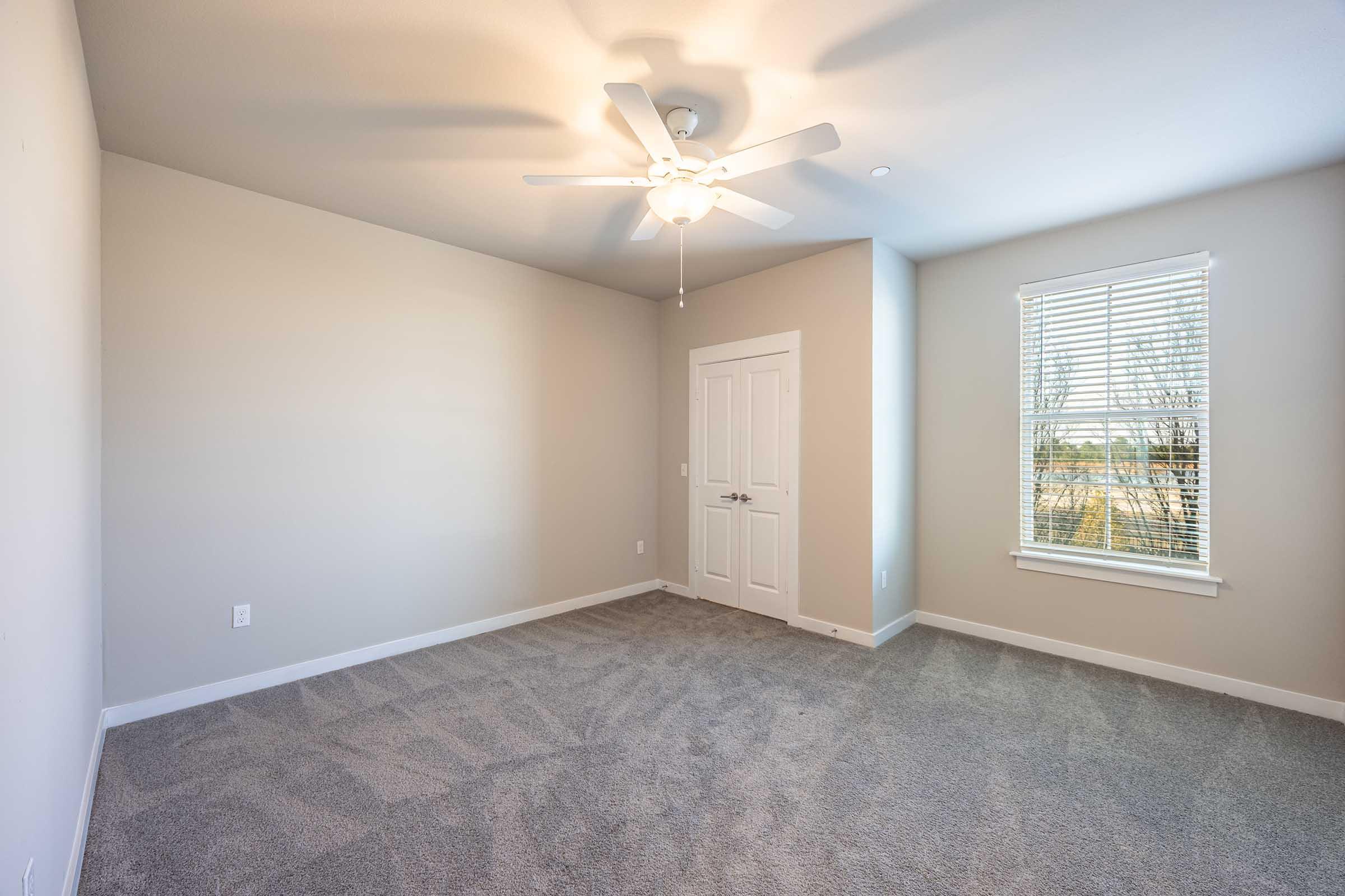 Empty room with light gray carpet, a ceiling fan, and soft beige walls. A window with blinds allows natural light to enter, and a white door is visible on the side. The room has a clean and spacious feel, with no furniture present.
