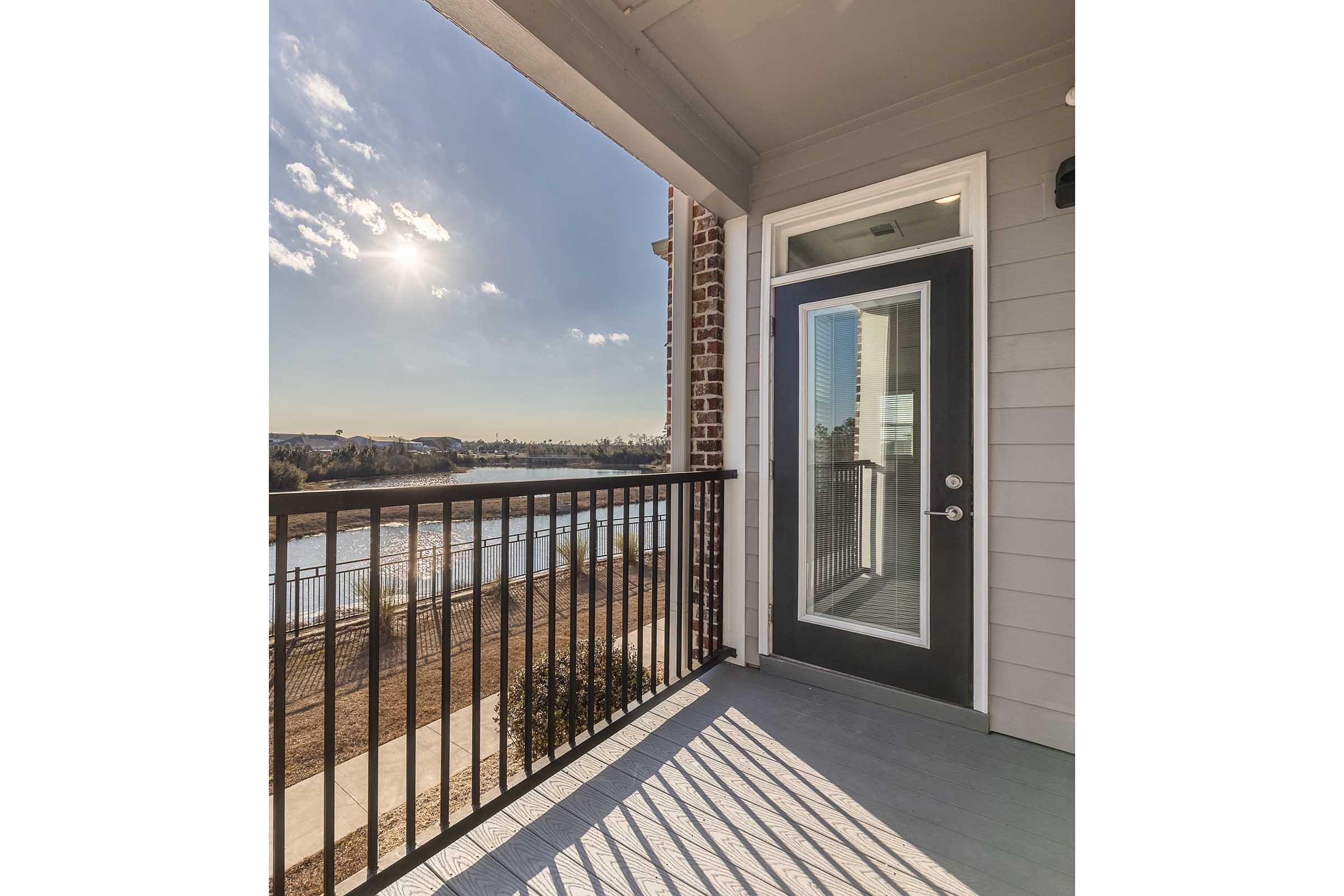 A view from a balcony featuring a door leading outside, with a railing and floor. In the background, there is a body of water reflecting the sky, and the sun is shining brightly, casting light and shadows. The scene conveys a serene outdoor atmosphere with a clear sky.