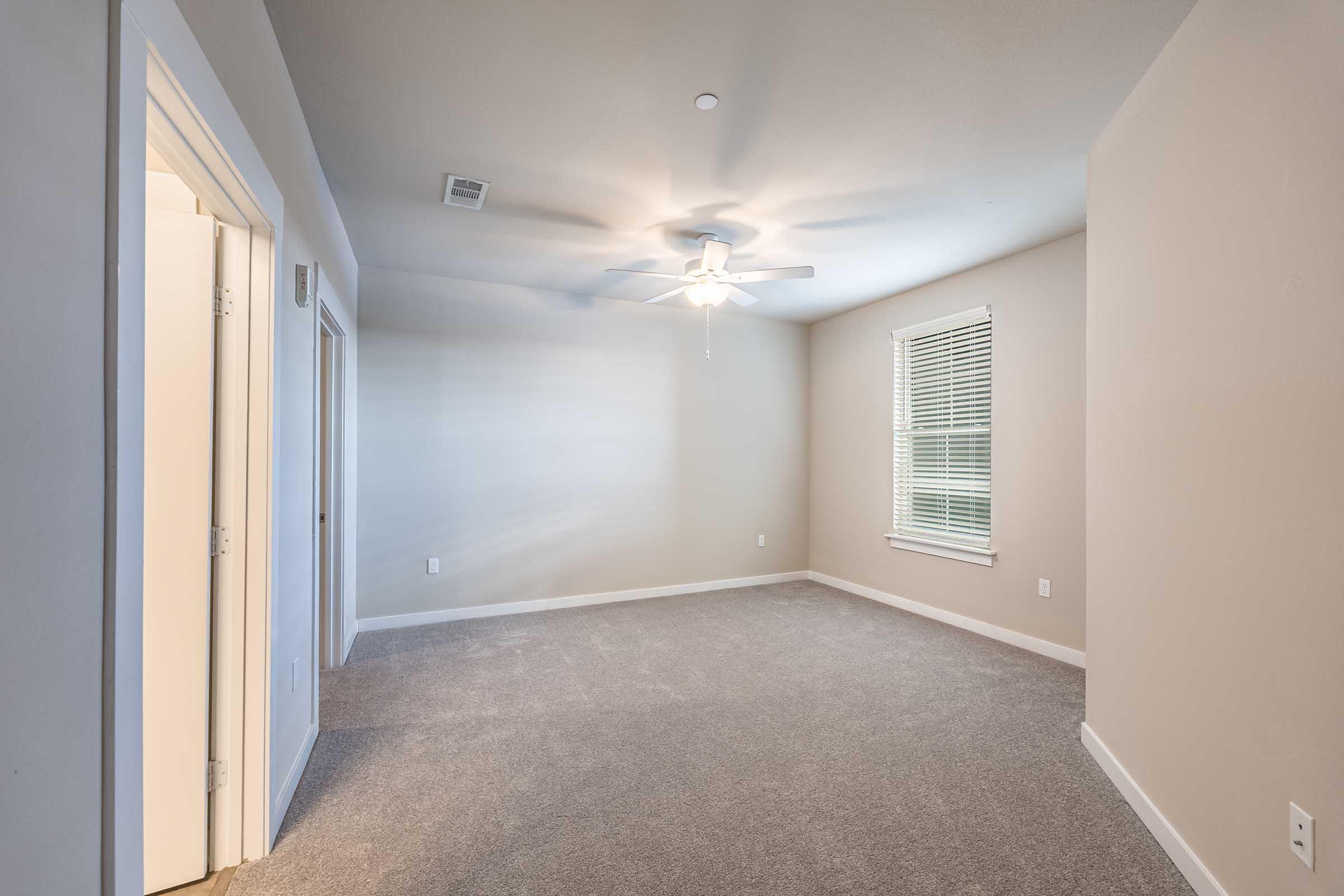An empty room featuring light-colored walls and carpeted flooring. A ceiling fan is installed, and there is a window with blinds allowing natural light. One wall has two doors, possibly leading to a closet or another space. The room appears spacious and clean, suitable for various purposes.