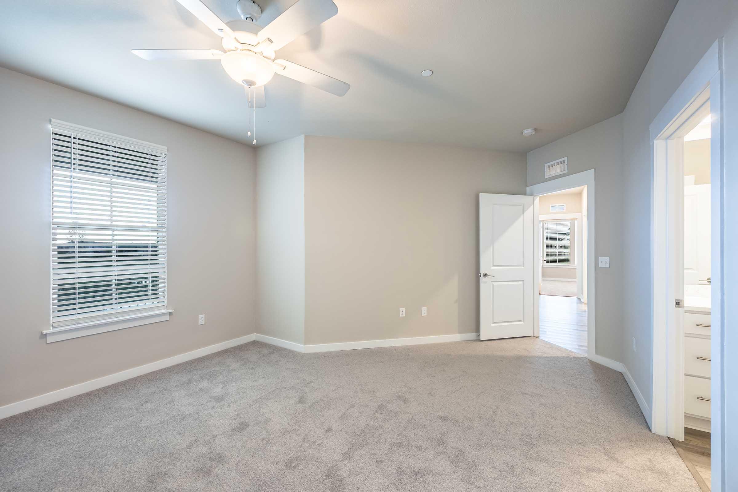 A spacious, lightly colored bedroom with a ceiling fan and carpeted floor. A window with horizontal blinds lets in natural light. An open door leads to another room, and the walls are painted in a soft neutral tone, creating a fresh and inviting atmosphere.