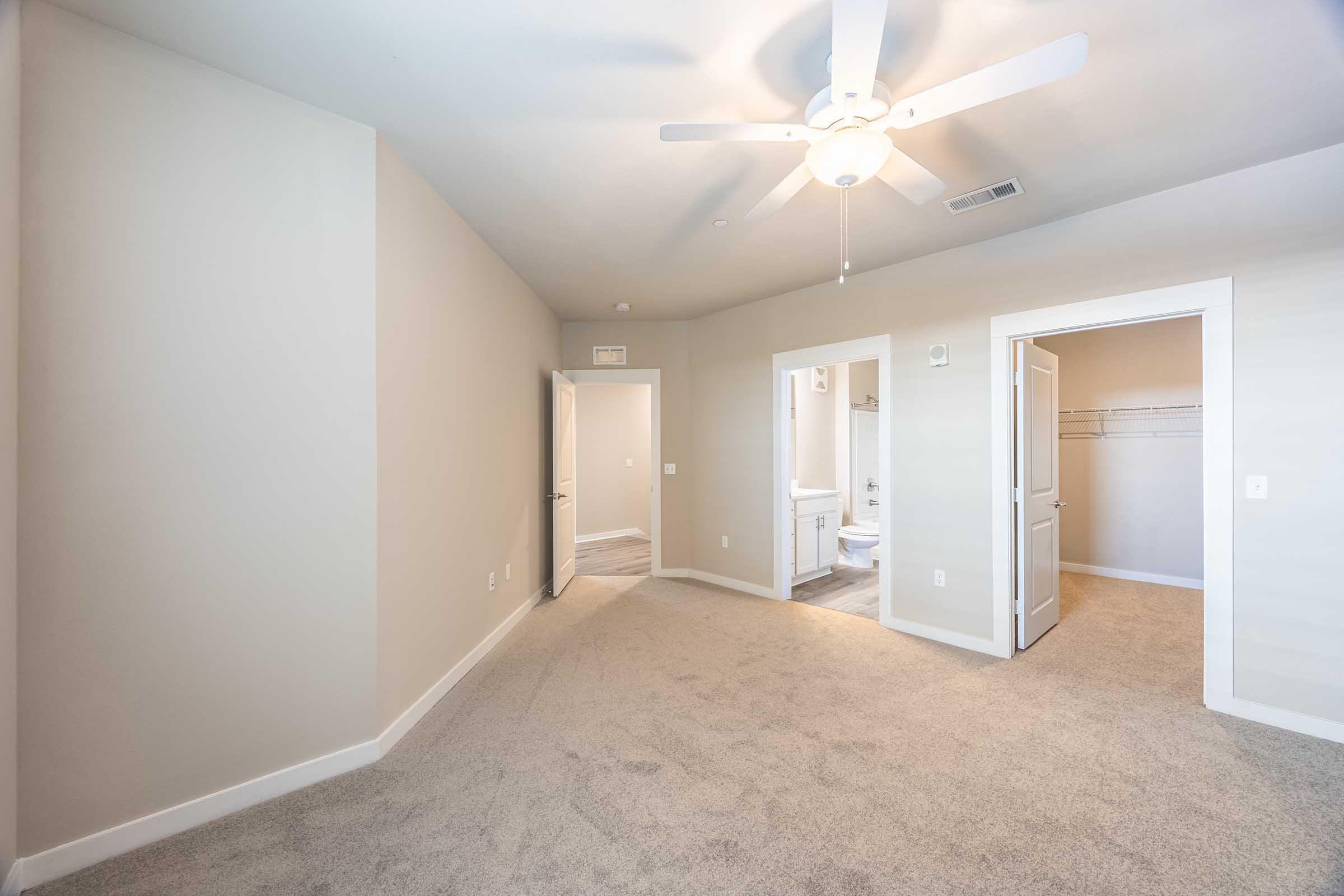A spacious, empty room with beige walls and carpet. It features a ceiling fan overhead and has two doorways leading to adjacent areas. One door opens to a bathroom area, while the other leads to a closet. Natural light brightens the space, creating an inviting atmosphere.