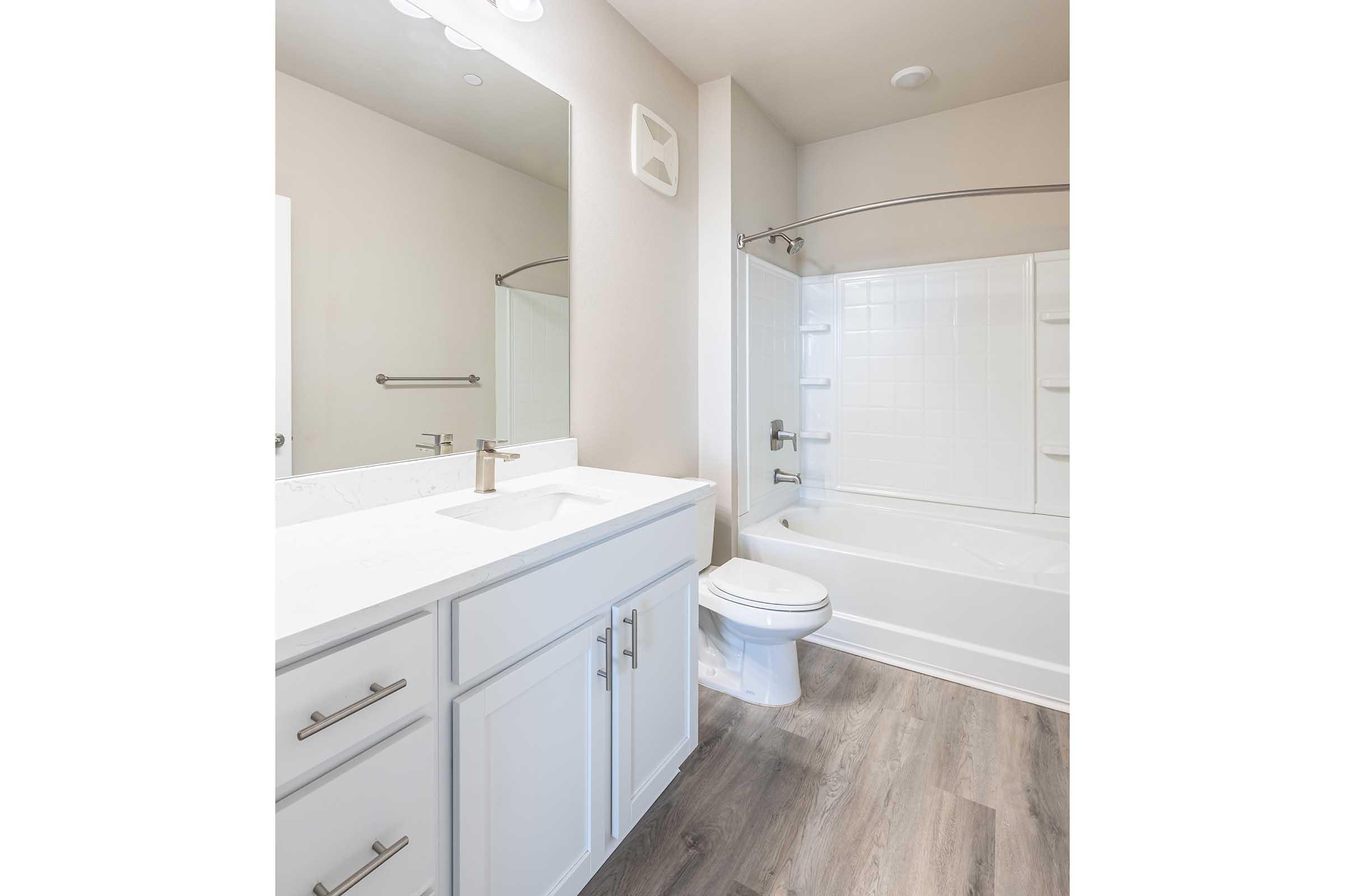 A modern bathroom featuring a white vanity with a sink, a large mirror above, and a bathtub with a shower. The walls are painted in a light color, and the flooring is a light wood laminate. The space is well-lit with overhead lighting.