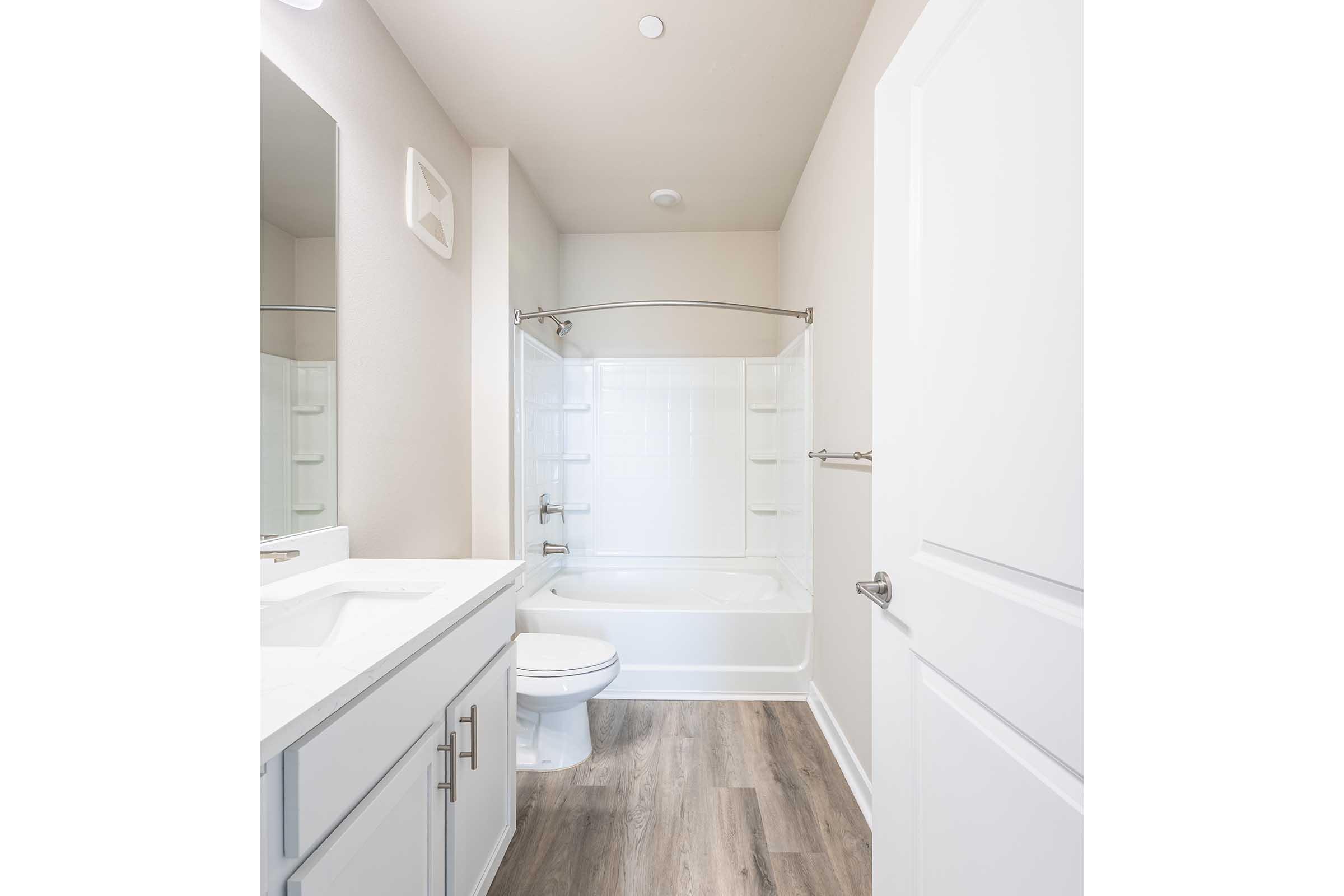 Modern bathroom featuring a white bathtub and shower combination, a vanity with a sink, a large mirror, and light-colored walls. The flooring is wood-like, adding warmth to the space. A towel rack is mounted on the wall, and there is ample natural light from a window.