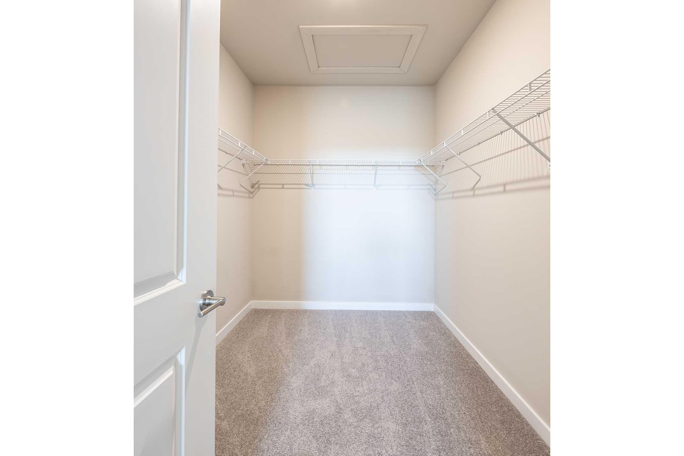 A spacious, empty closet with light-colored walls and plush carpet. It features two wire shelves mounted on the walls, providing storage options, and a white door is partially open, revealing the interior space. The room is well-lit and unoccupied, emphasizing its potential for organization.