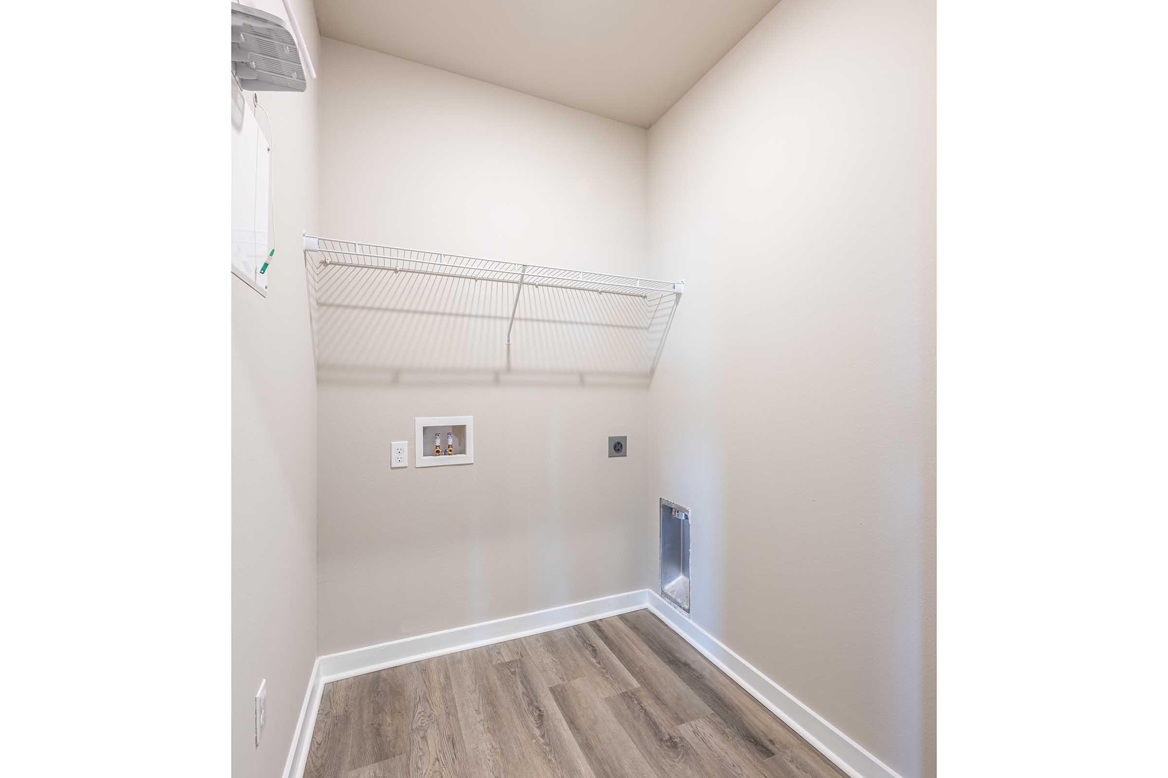 A laundry room with a light-colored wall, a wire shelf mounted above, and a utility area. The floor is covered in light wood-like laminate. There are electrical outlets and a vent on the wall, but the space is empty and uncluttered, giving a clean and modern appearance.