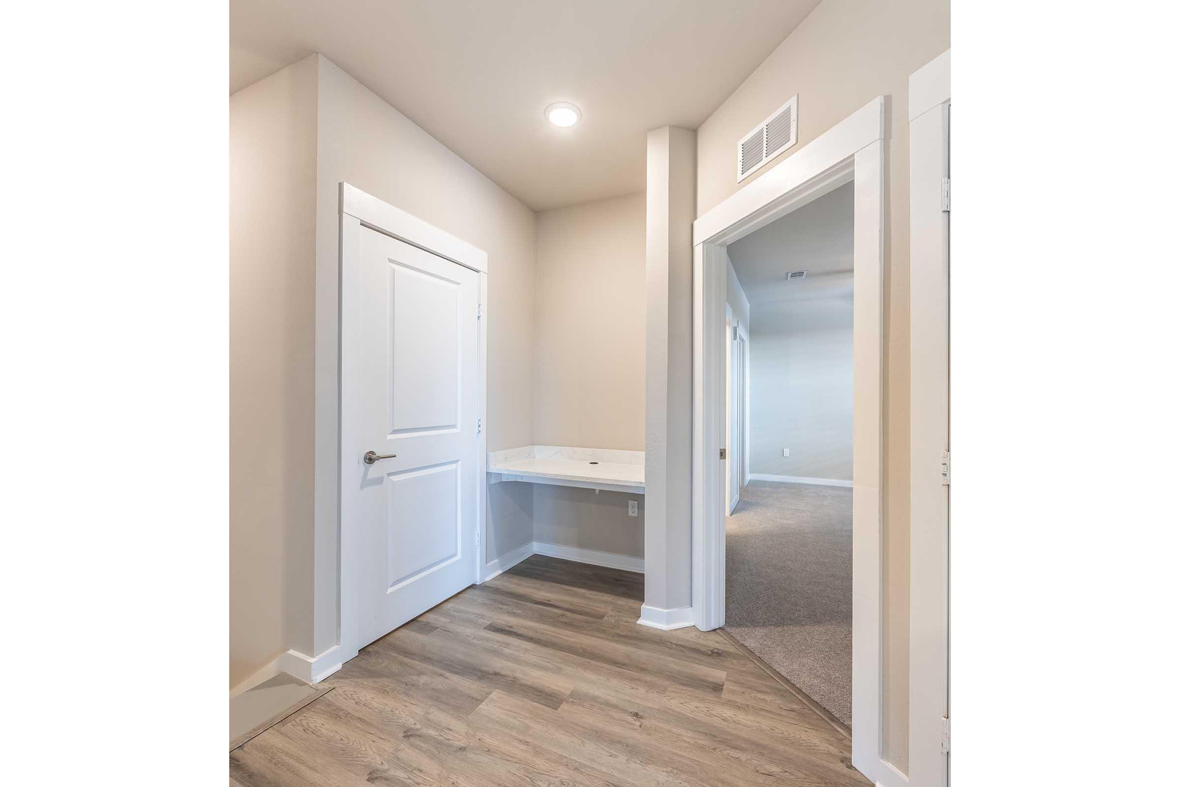 Interior view of a hallway featuring a doorway with a white door on the left, a small countertop area against a light-colored wall, and another doorway on the right. The flooring is a wooden laminate, and the space is illuminated by a ceiling light, creating a bright and open feel.