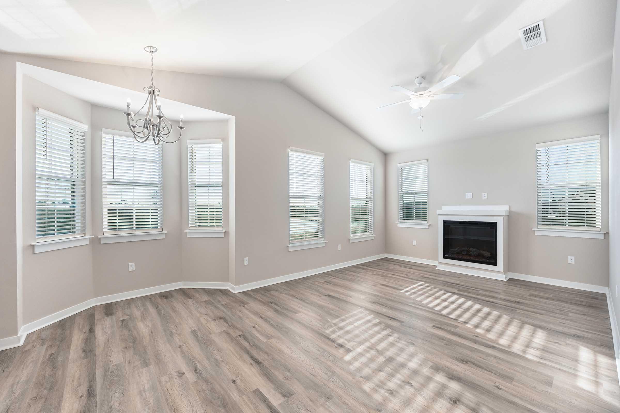 Spacious living room with vaulted ceiling, large windows with blinds, and light-colored walls. Features a decorative chandelier, modern ceiling fan, and a corner fireplace. The flooring is light wood, enhancing the airy atmosphere. Natural light filters in, creating a warm and inviting space.