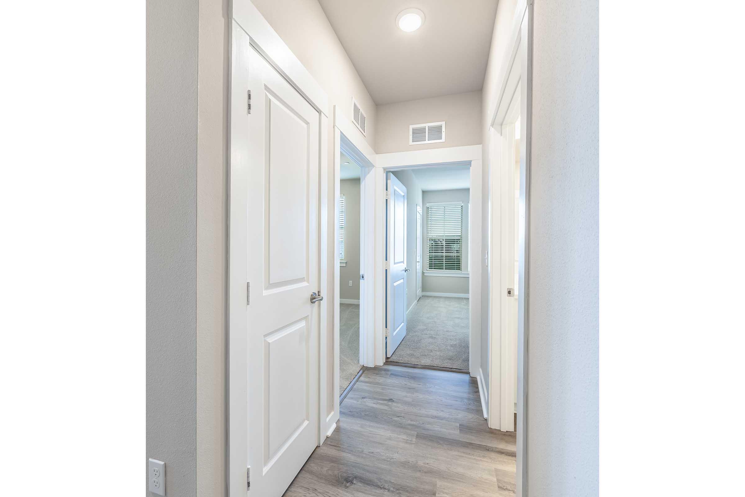 A well-lit hallway featuring light-colored walls and wooden flooring. Multiple doorways lead to adjacent rooms, with one doorway at the end opening into a carpeted space. Natural light filters through the windows on the left, enhancing the inviting atmosphere.