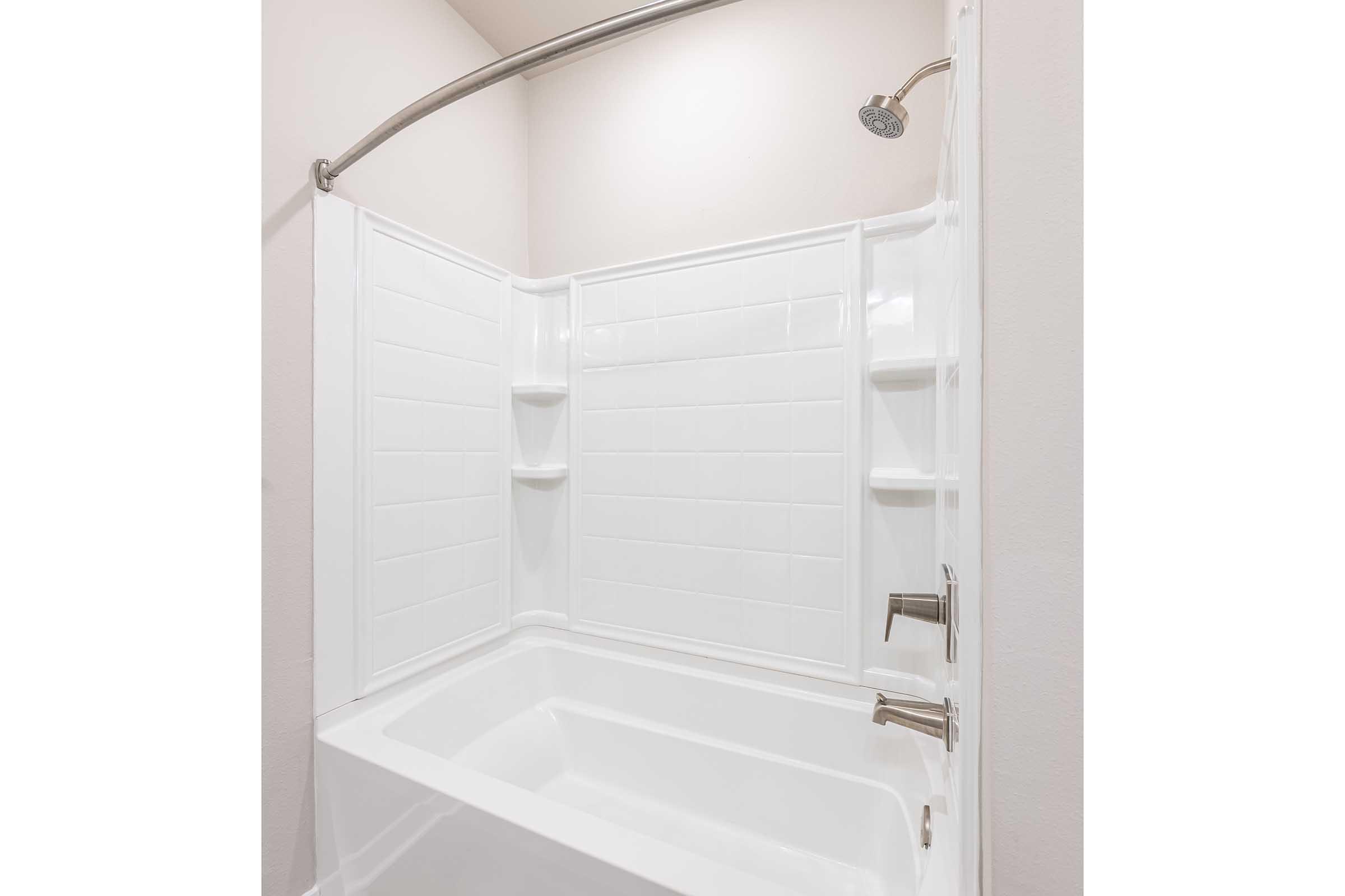 A clean, modern bathroom featuring a white bathtub with a curved shower rod and a showerhead above. The walls are tiled with white panels, and there are built-in shelves on one side of the tub for toiletries. The overall aesthetic is bright and minimalist.