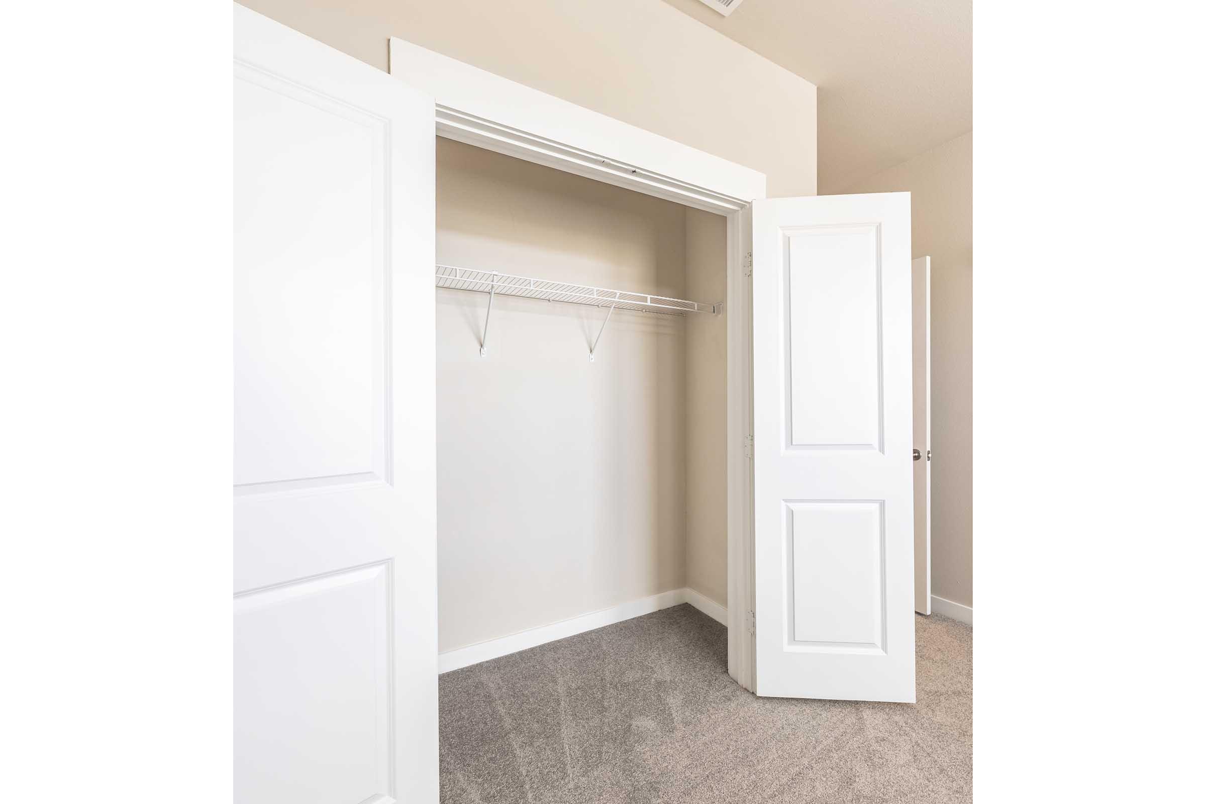 A clean, empty closet with white double doors open, revealing beige walls and light carpet. The closet features a simple wire shelf and two hanging rods, indicating it is ready for storage or organization. The overall space is well-lit and tidy.