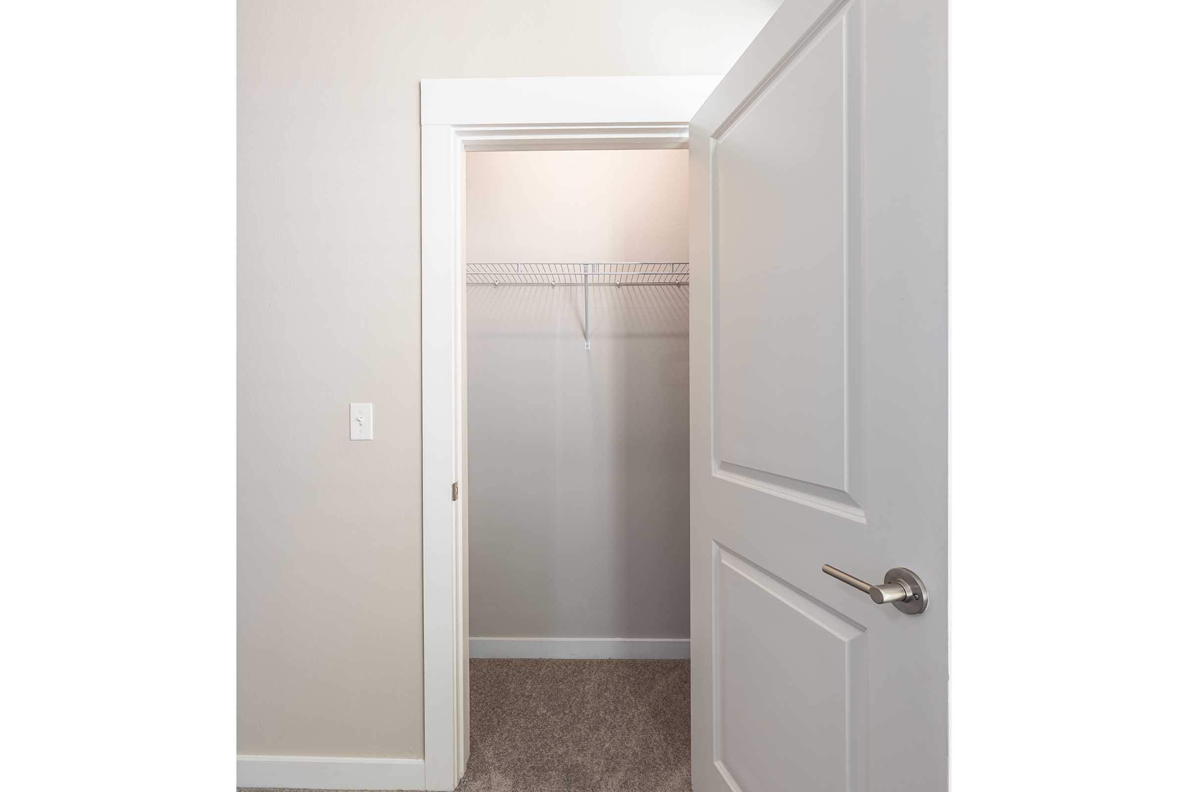 An open closet with a white door, featuring a wire shelf along the top. The walls are painted a light color, and the floor is covered with a soft, neutral carpet. The space is empty and minimalistic, providing a clean and organized look.