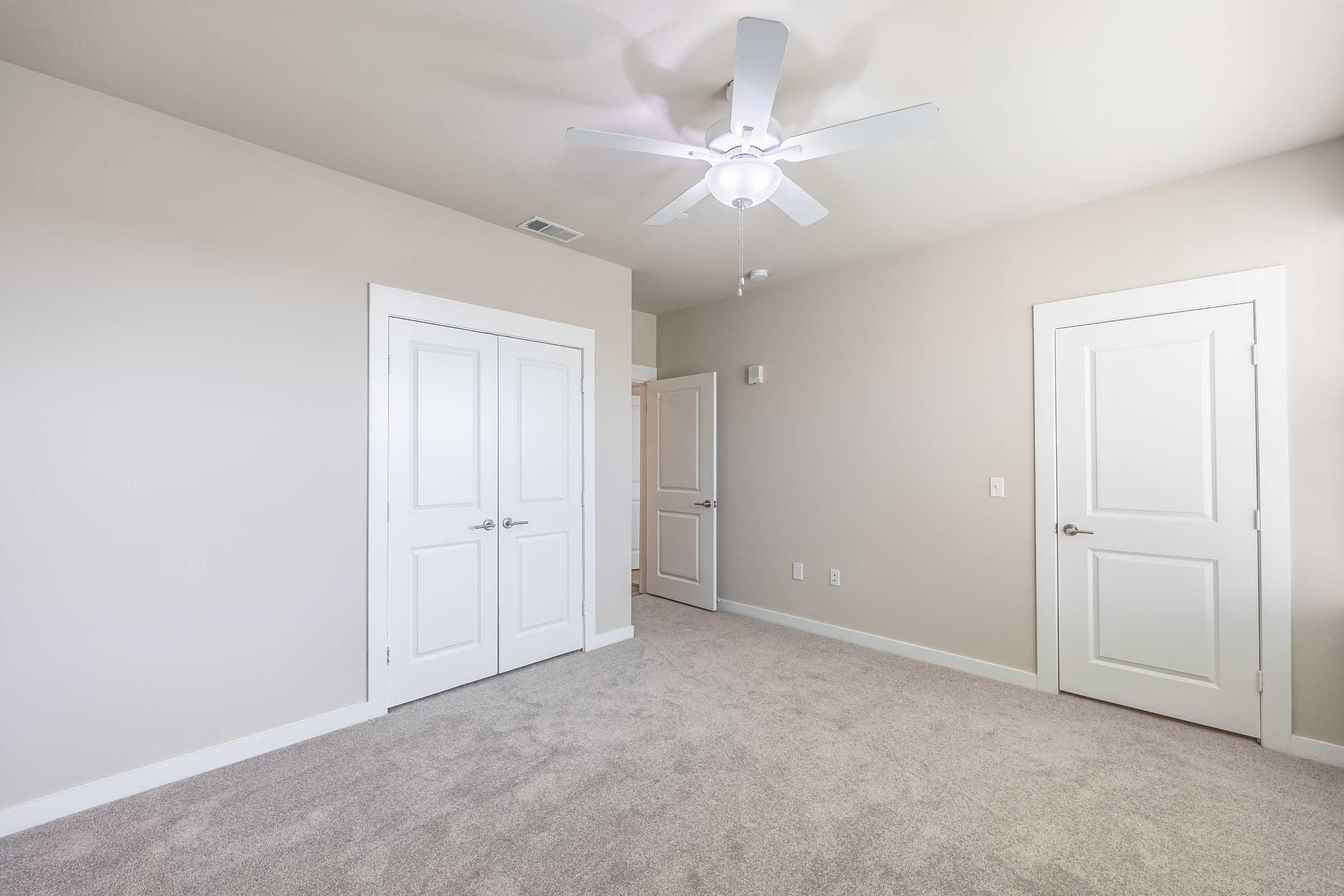 Bright, empty room with light beige walls and carpet. A white ceiling fan is overhead, and there are two closed white doors on the left side leading to other spaces. Soft natural light fills the room, creating a clean and airy atmosphere.