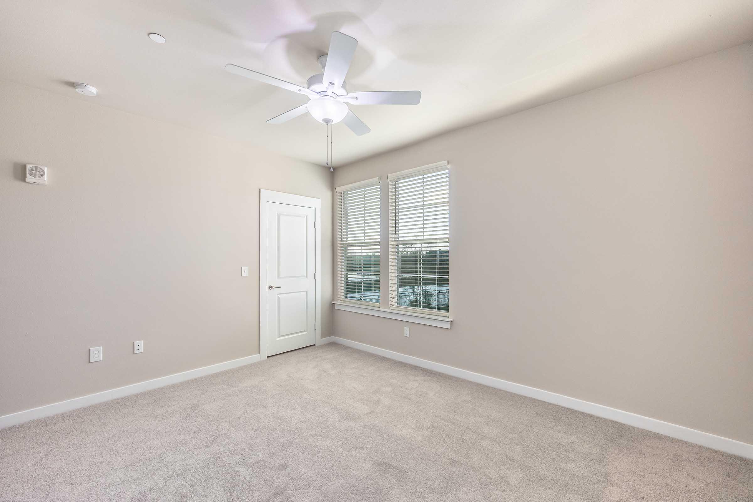 Empty room featuring light beige walls, a ceiling fan with a light fixture, and large windows with white blinds. The floor is covered in soft carpet, and there is a door leading to another room. The space is well-lit and showcases a minimalist design.