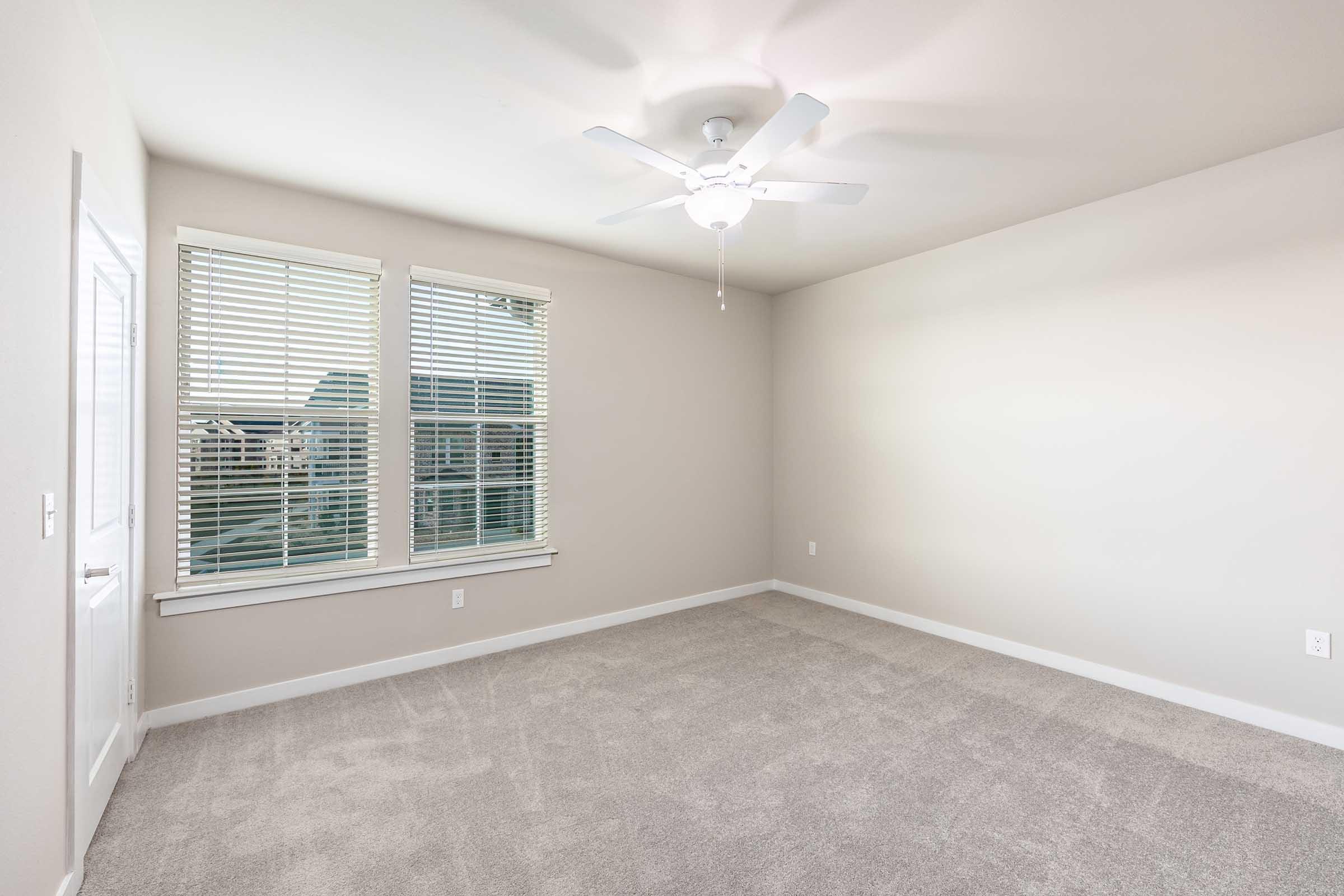 Empty room with light-colored walls and carpeted floor. A ceiling fan is mounted in the center, and there are two windows with white blinds on one wall, allowing natural light to fill the space. The opposite wall features a door, suggesting entry to another area.
