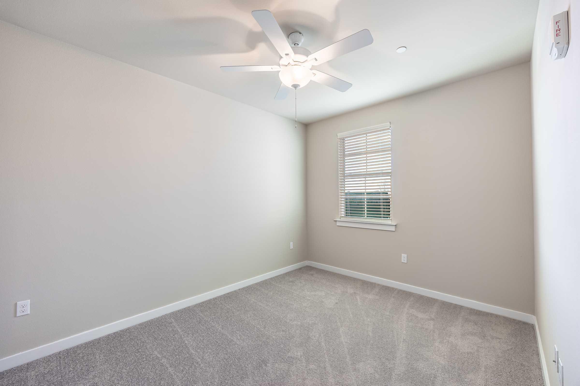 Empty room with light gray carpet, off-white walls, and a white ceiling fan. A window with blinds allows natural light to enter, and there are no furnishings or decorations. The room has one wall-mounted electrical outlet and a smoke detector.