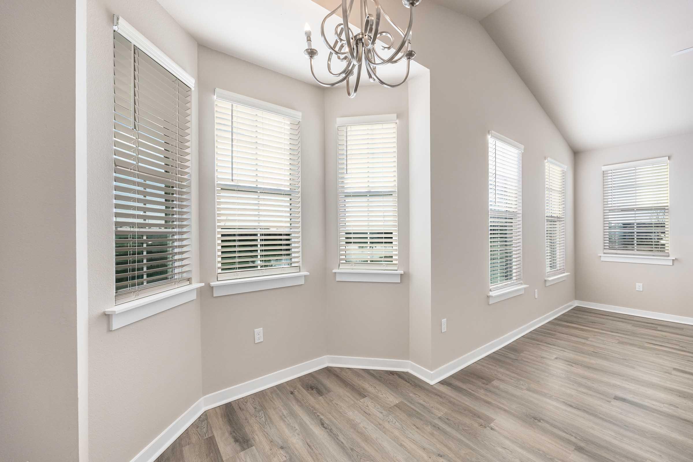Bright and airy living space featuring multiple windows with blinds, allowing natural light to filter in. The walls are painted in a soft neutral color, and the flooring is a light wood. A modern chandelier hangs from the ceiling, complementing the clean and contemporary design of the room.
