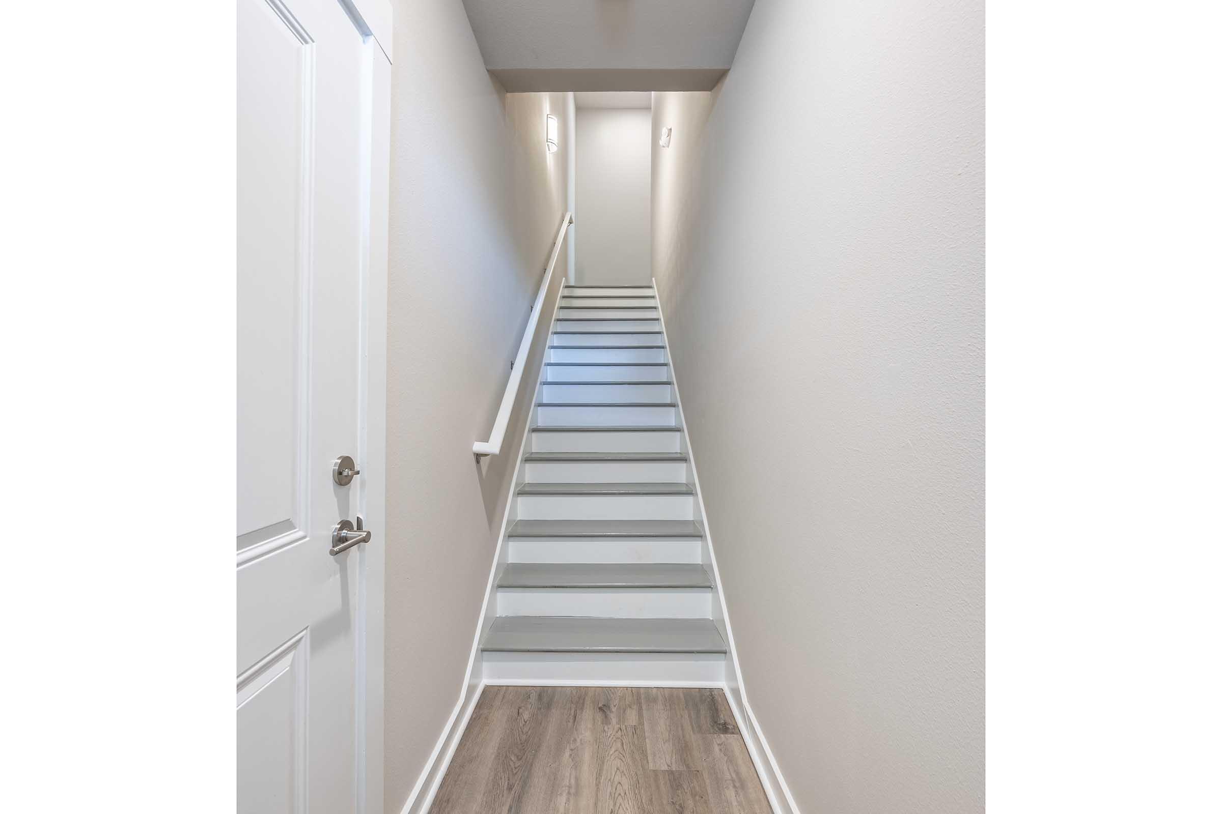 Bright, clean stairwell leading upward. The stairs are painted white, and the walls are a light beige. A door is visible on the left side, and soft lighting illuminates the hallway above. The floor features a wood-like finish, creating a modern and inviting atmosphere.