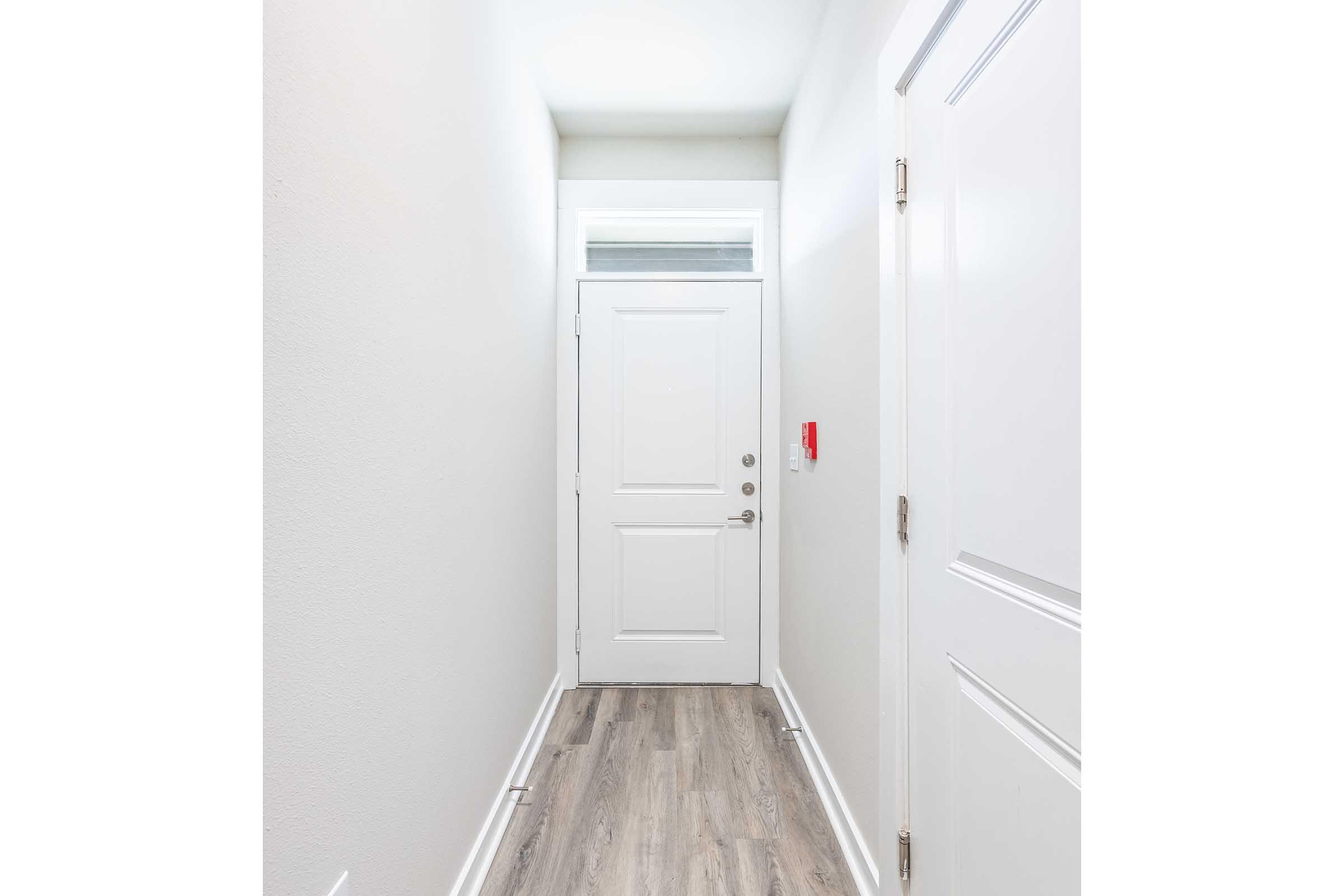 Narrow hallway featuring a white front door at the end, light gray walls, and light wood flooring. A small window above the door provides natural light, and there is a red security system panel on the wall to the right. The overall atmosphere is bright and modern.