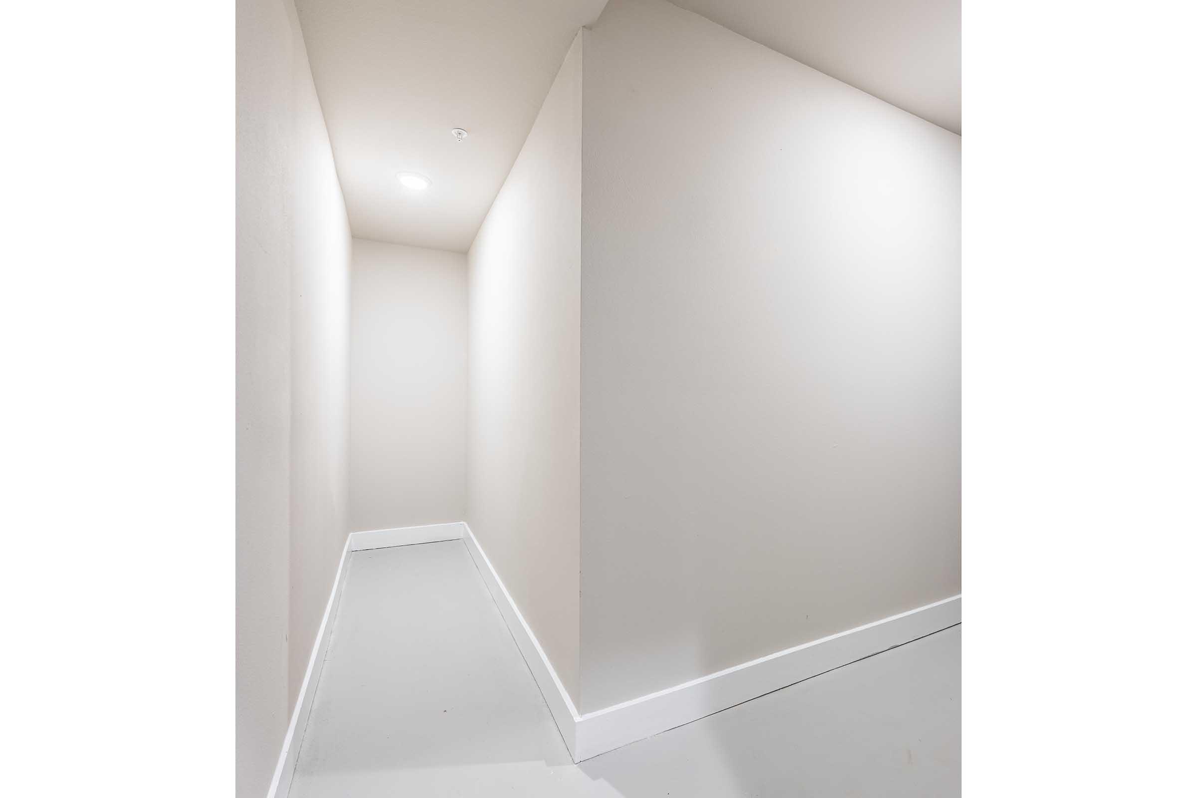 A narrow, well-lit hallway with plain white walls and a smooth gray floor. The space is devoid of decorations or furnishings, creating a minimalist atmosphere. The lighting is bright, enhancing the clean and simple design of the corridor.