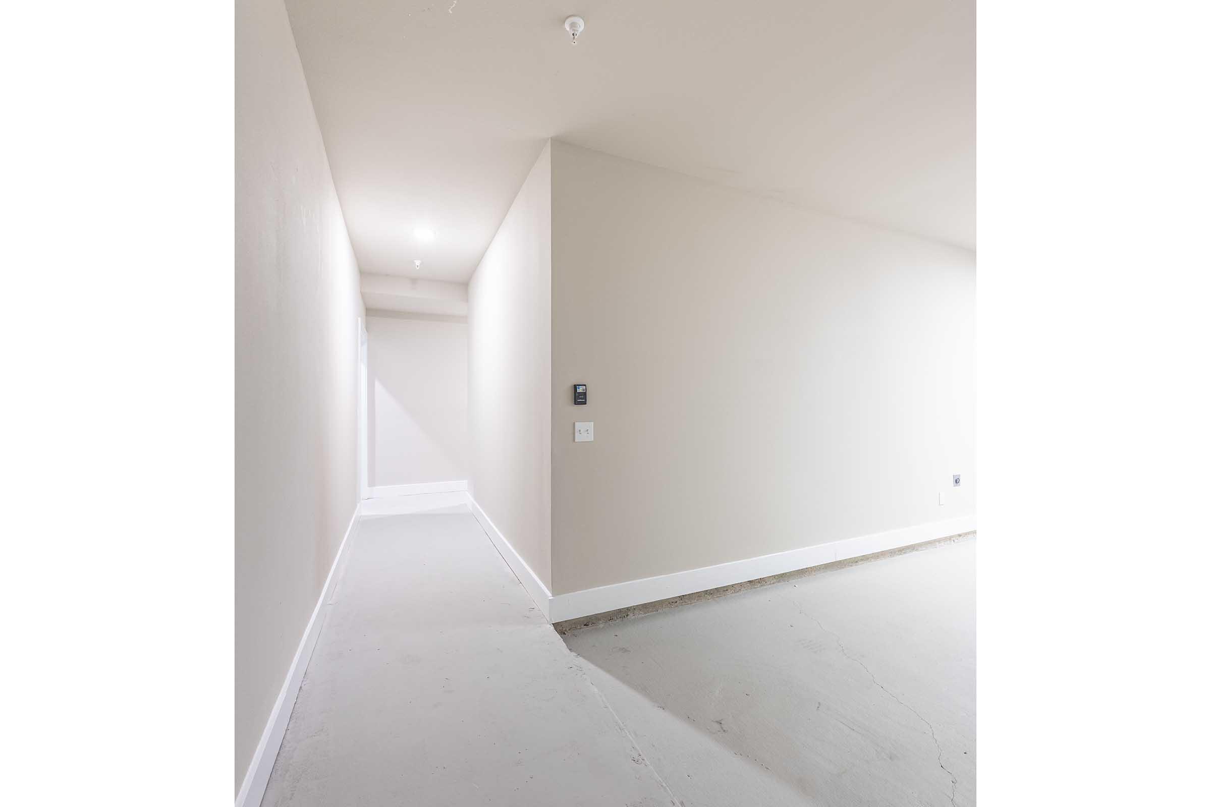 A long, empty hallway with light beige walls and a concrete floor. The hallway leads into a bright, unremarkable space at the end. A light switch is visible on the wall, and the overall atmosphere is minimalistic and freshly painted.