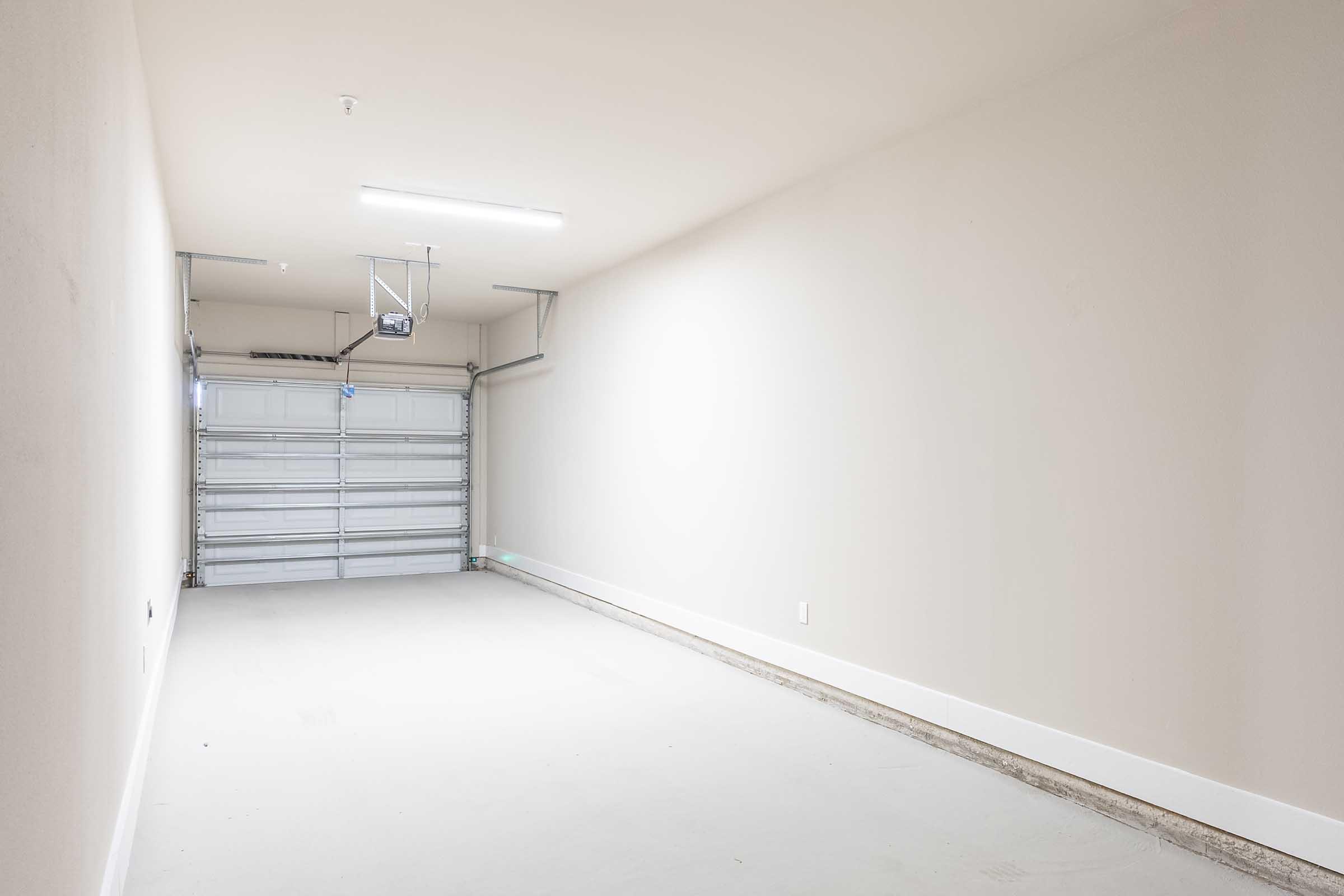 A spacious, empty garage with a clean concrete floor and a faintly illuminated ceiling. There is a closed sectional garage door on one side. The walls are painted a light, neutral color, creating a bright and unobtrusive atmosphere.