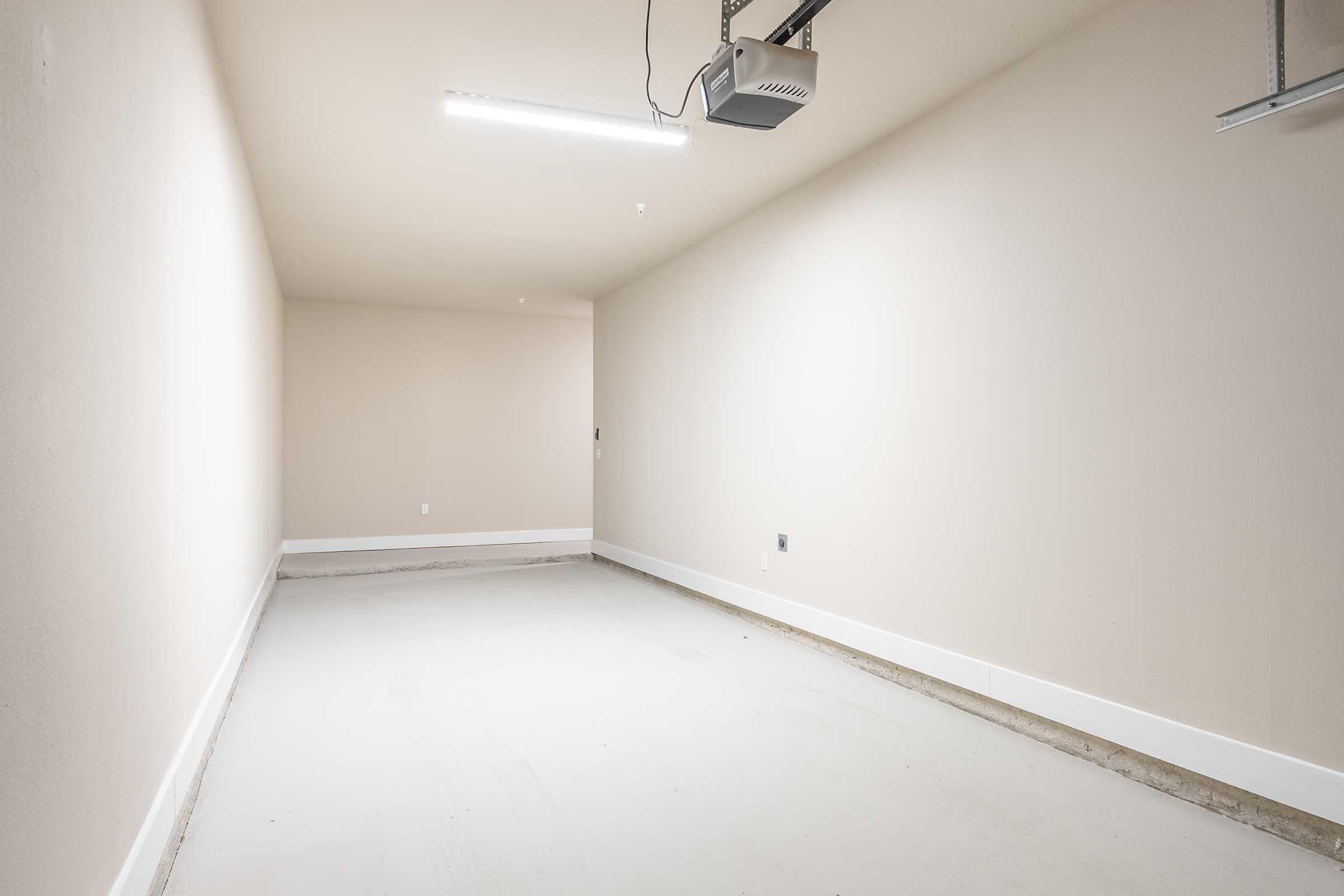 A spacious, empty garage with light gray walls and concrete flooring. There is a ceiling-mounted garage door opener visible, and the room is well-lit with fluorescent lighting. The overall appearance is clean and uncluttered, providing a blank canvas for storage or modification.