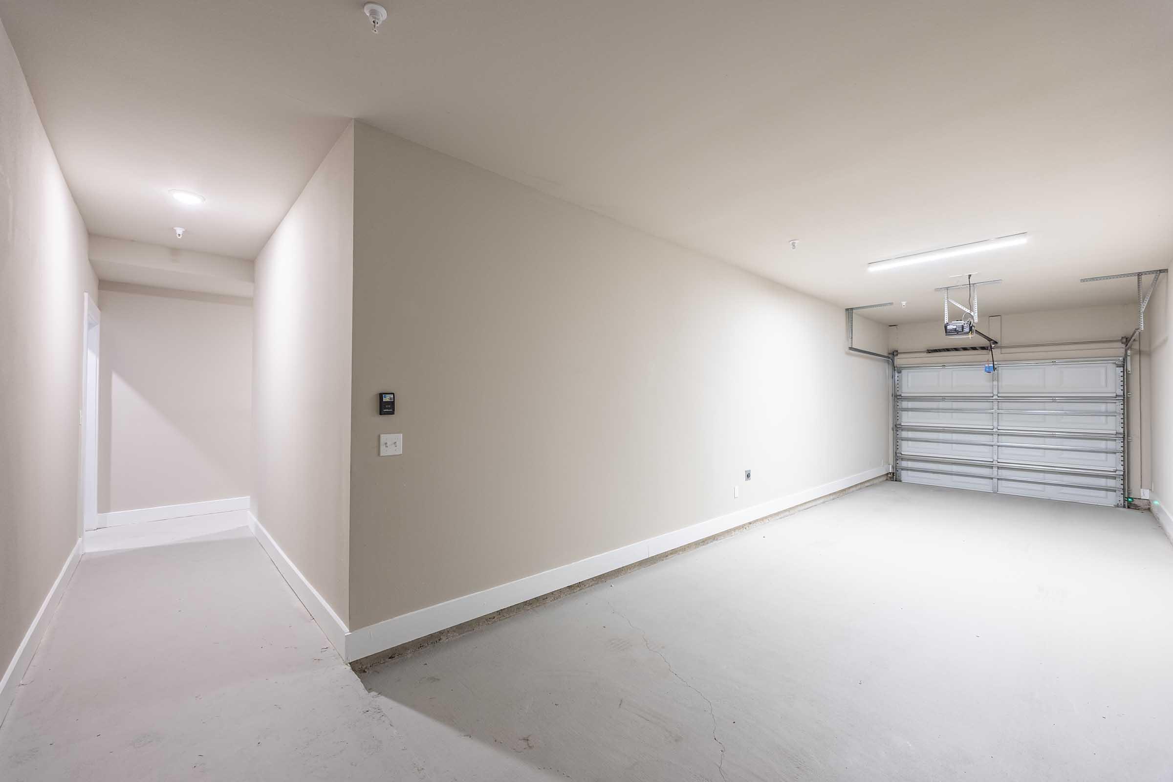 Empty garage interior with a light beige wall and a concrete floor. A closed garage door is visible at the far end, and there is a hallway leading to a door on the left. The space is well-lit with overhead lighting, creating a clean and open atmosphere.