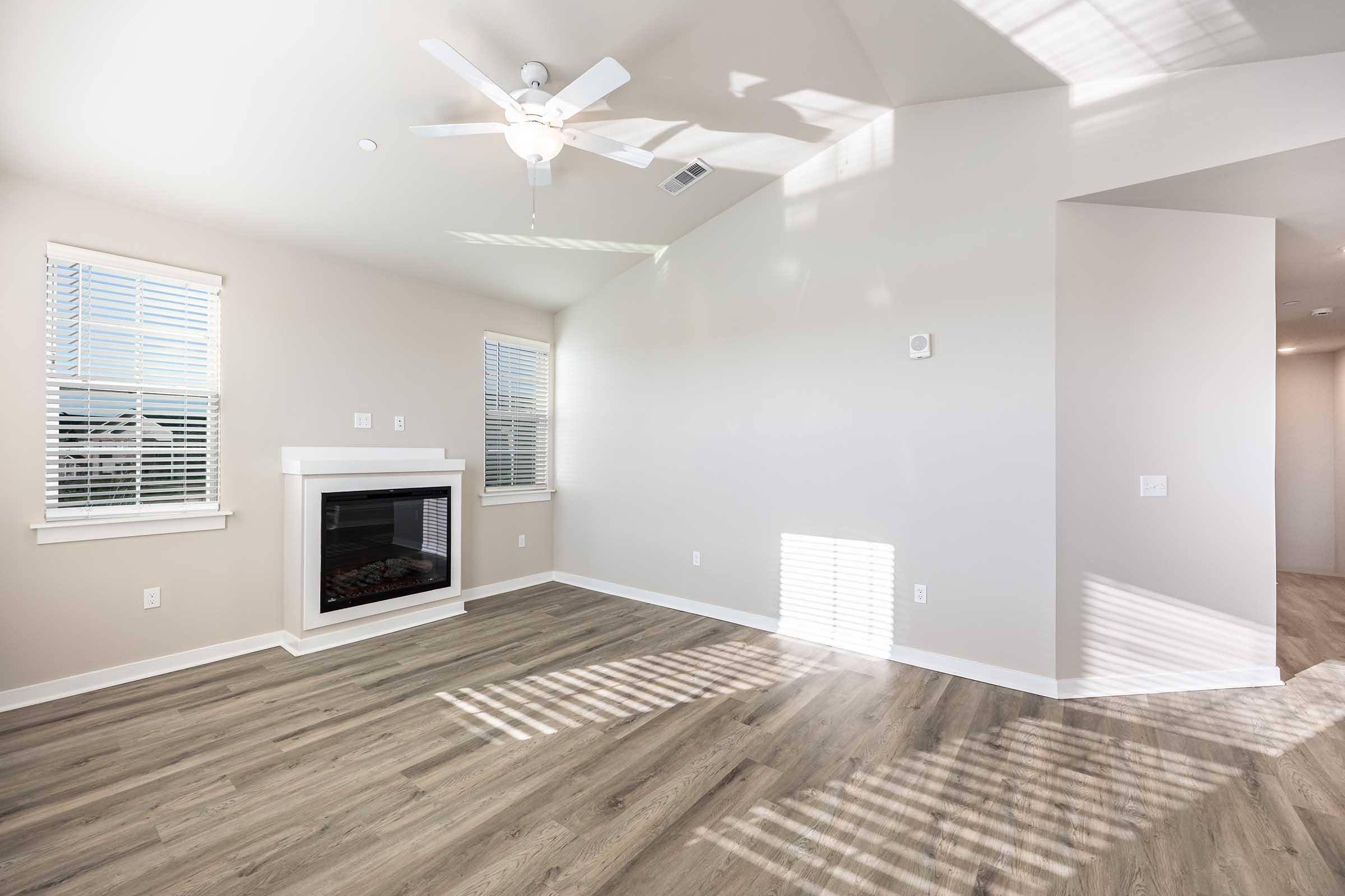 Bright and spacious living room featuring a ceiling fan, large windows with natural light, and light-colored walls. A sleek electric fireplace is situated on one wall, complementing the modern flooring. The area is free of furniture, emphasizing its open layout.