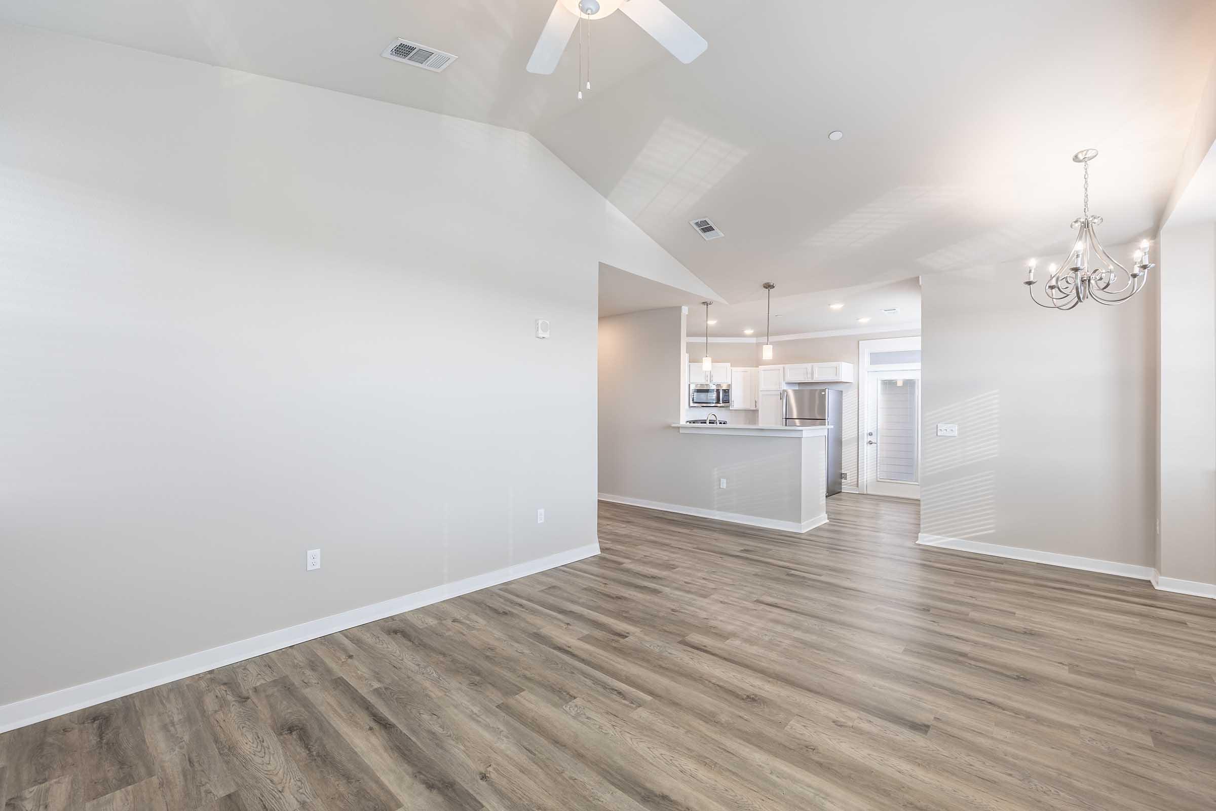 A spacious, newly built interior view of a living area featuring light-colored walls, modern light fixtures, and wood-style flooring. In the background, an open kitchen with stainless steel appliances is visible, along with a ceiling fan and large windows allowing natural light.