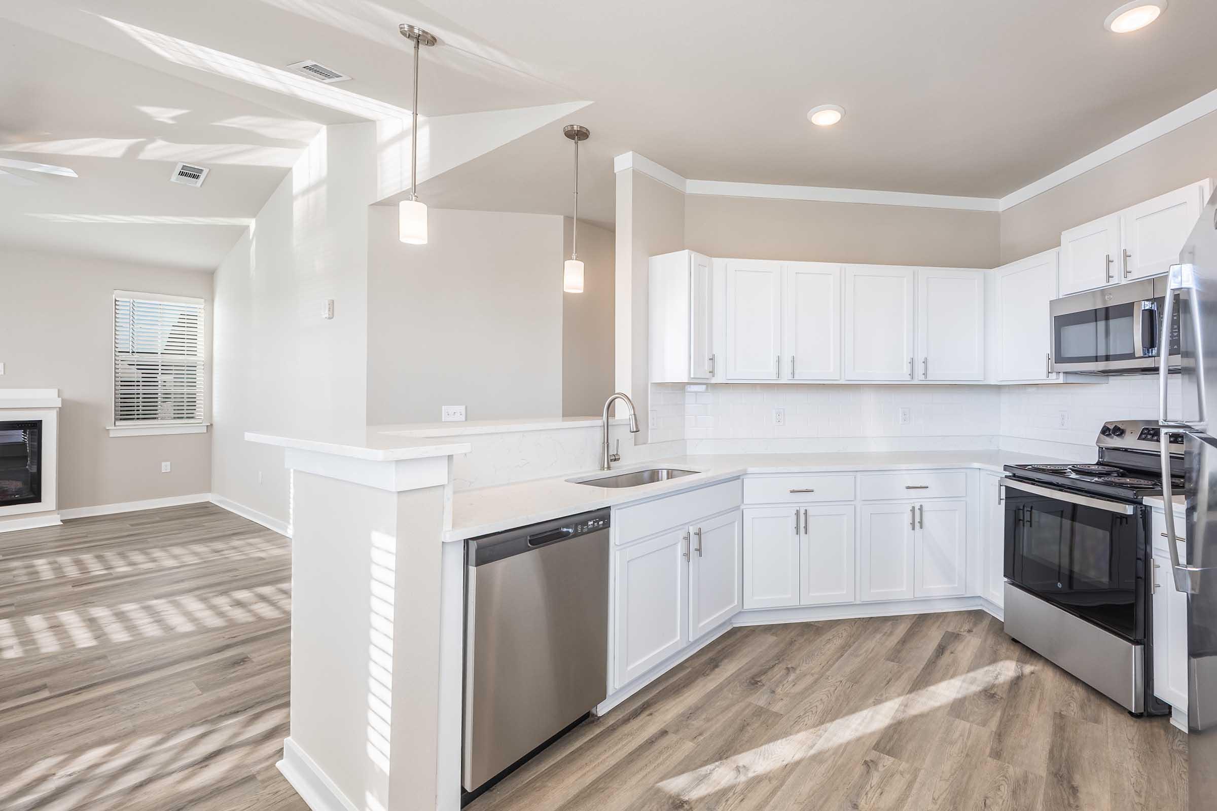 Bright, modern kitchen featuring white cabinetry, stainless steel appliances, and a spacious layout. Includes a double sink, dishwasher, and an open design with natural light streaming through windows. Light wood-style flooring complements the contemporary style.