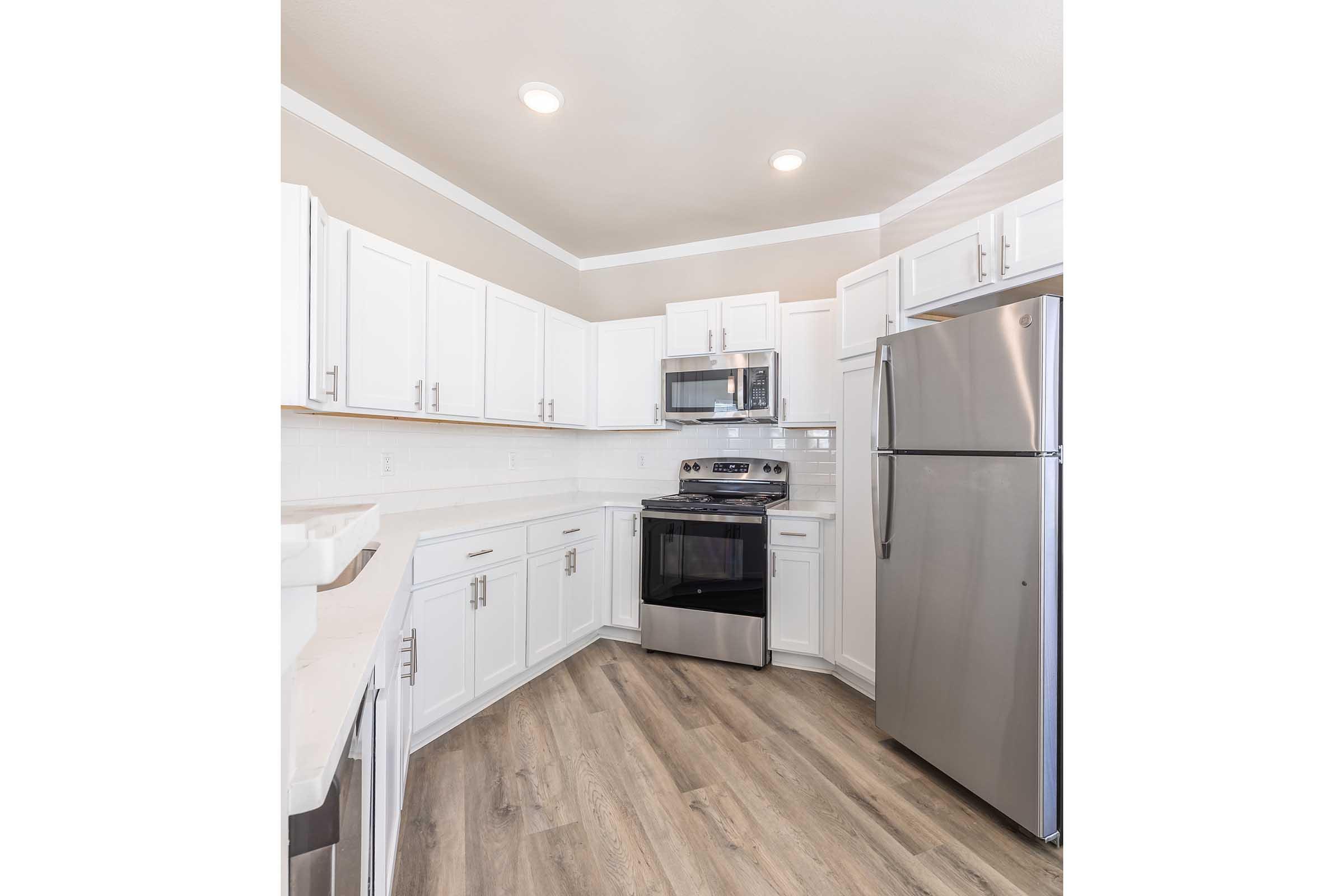 Modern kitchen featuring white cabinetry, stainless steel appliances, and light wood flooring. The space includes a silver refrigerator, a built-in microwave, and an oven with a smooth top range. Soft, neutral wall colors enhance the airy feel of the room, complemented by bright ceiling lights.