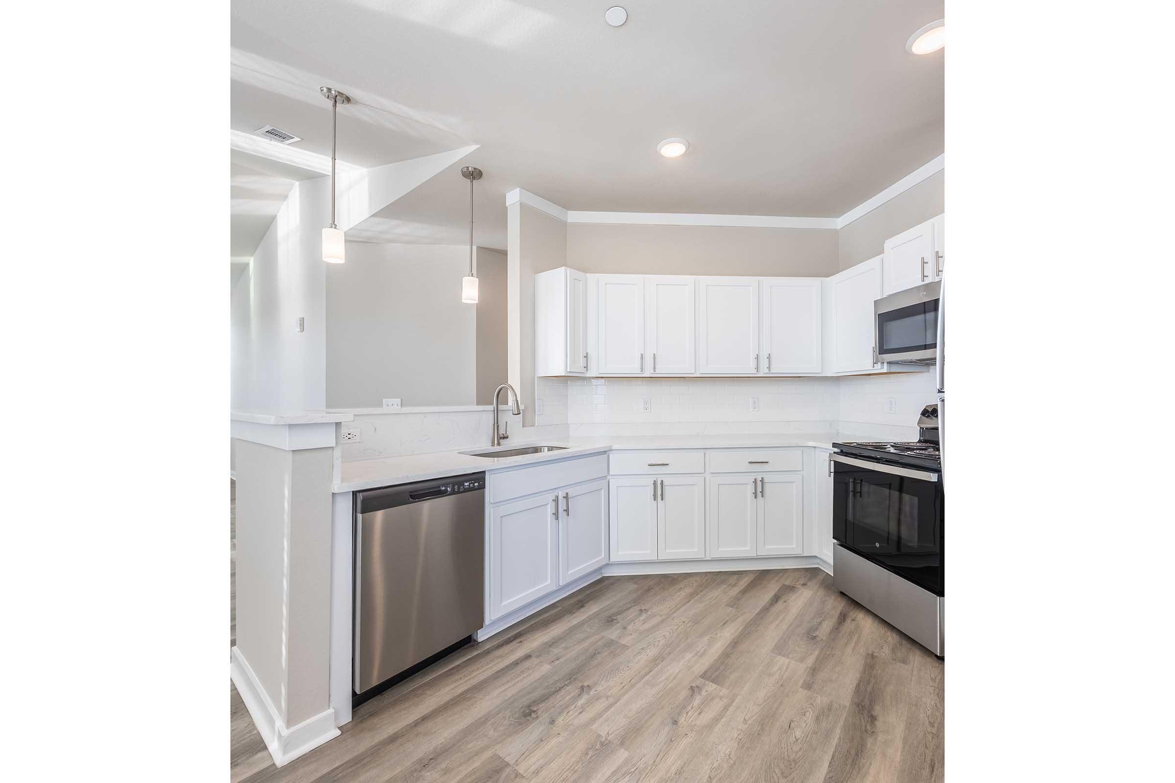 Modern kitchen featuring white cabinetry, stainless steel appliances, and a grey countertop. Includes a dishwasher, microwave, and stove, with pendant lighting above the counter. The floor is a light wood finish, creating a bright and inviting space.