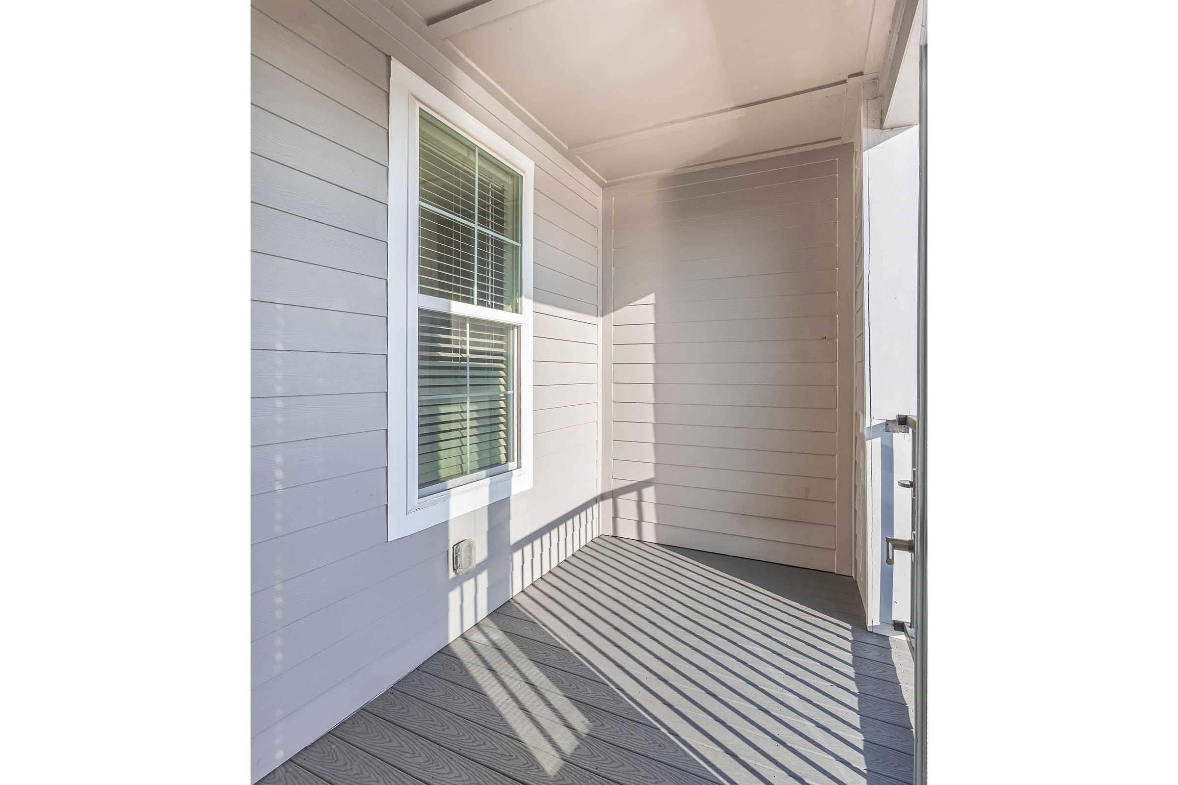 A well-lit covered porch featuring light gray walls and a wooden floor. A window on the left allows natural light to flood the space, creating distinct shadows on the floor. The overall ambiance is fresh and inviting, suitable for relaxation.