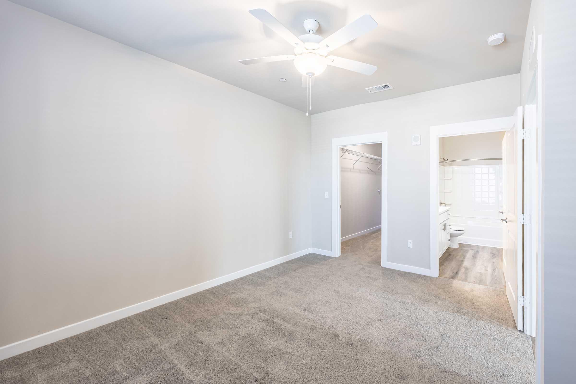 A spacious, empty room with light gray walls and plush carpet. A ceiling fan is installed, and there are two doorways: one leading to a bathroom with a light-colored tile and another to a closet with shelving. Natural light fills the space, creating a bright and airy atmosphere.