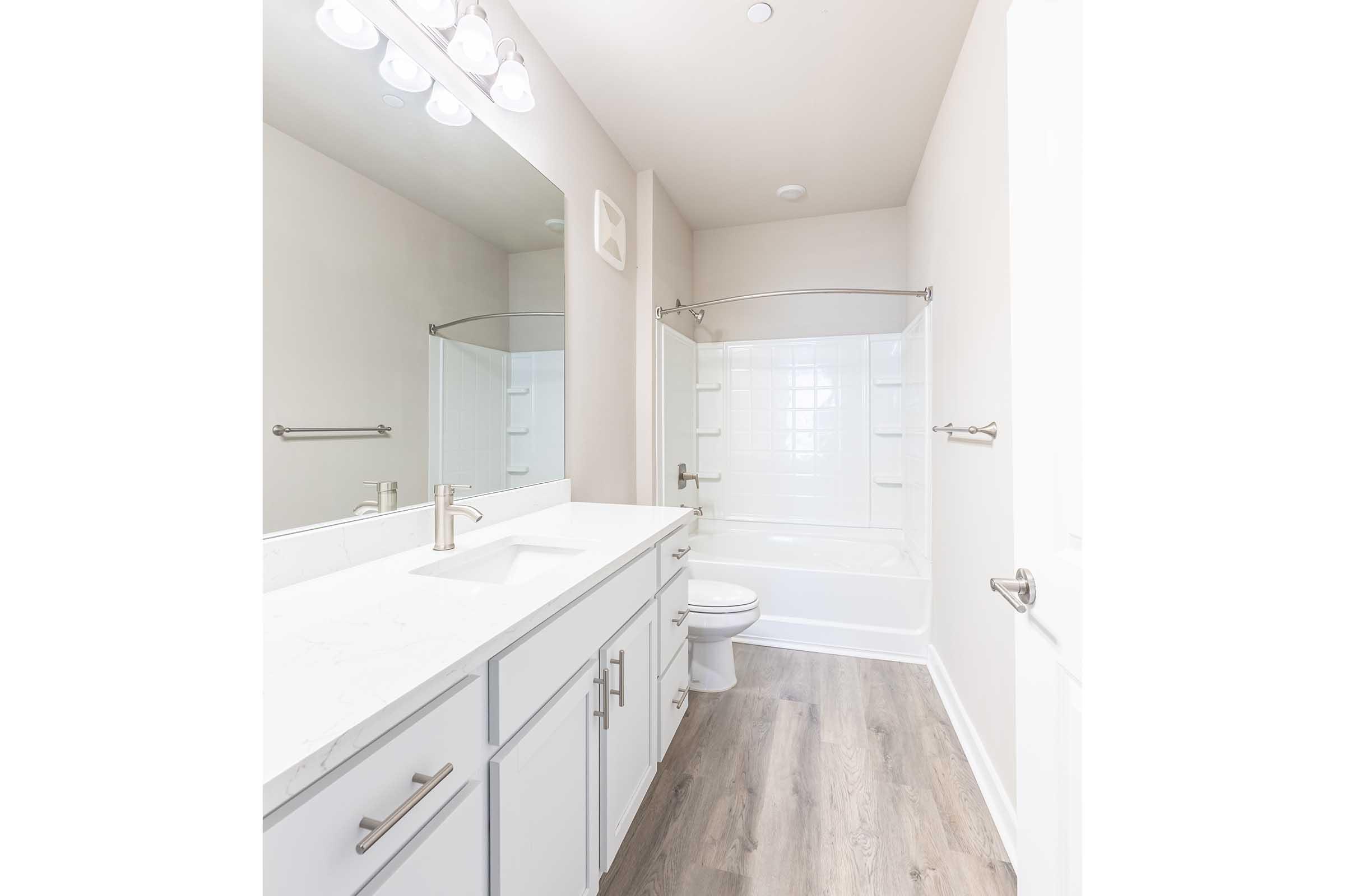 Modern bathroom featuring a white vanity with a marble countertop, a large mirror with three light fixtures above, and a clean, glass-enclosed shower. The flooring is light wood laminate, and there is a white bathtub with a shower curtain. The walls are painted a soft neutral color.