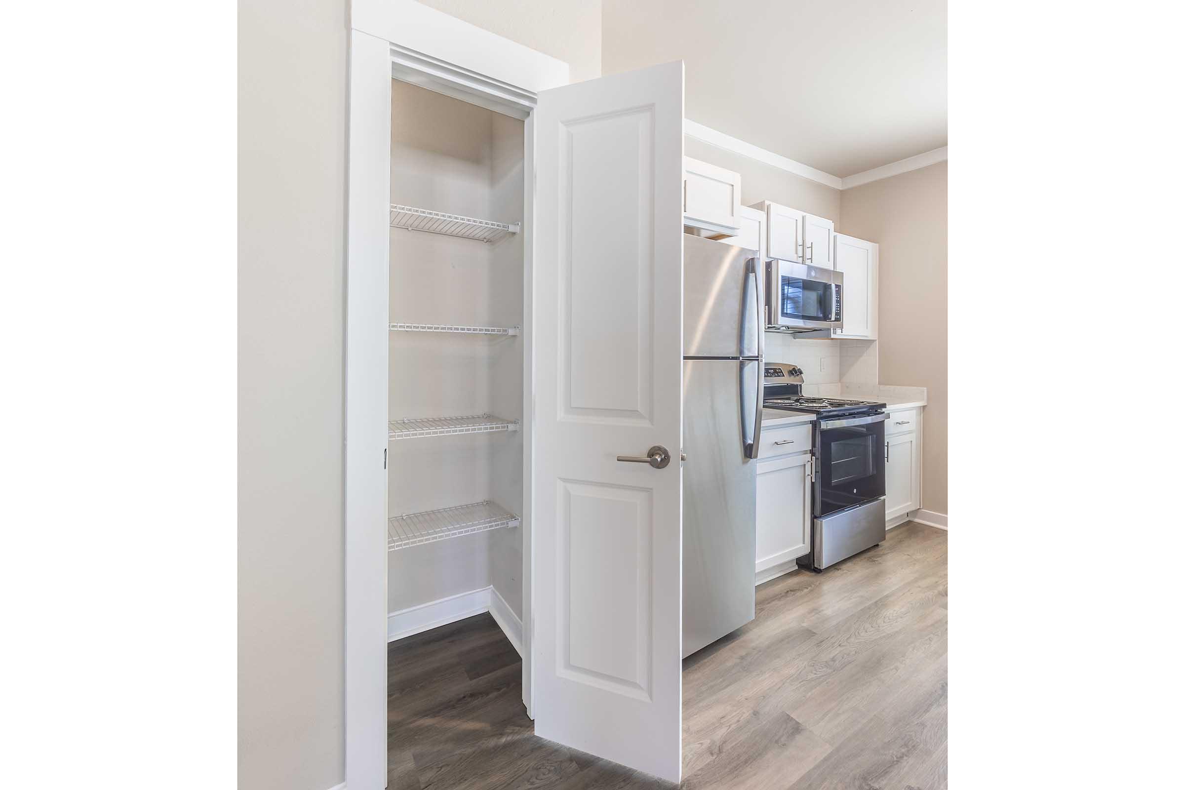 A clean and modern kitchen featuring white cabinetry, stainless steel appliances, and a pantry door slightly open, revealing empty shelves. The flooring is a light wood laminate, and the walls are painted a soft neutral color, creating an inviting atmosphere.