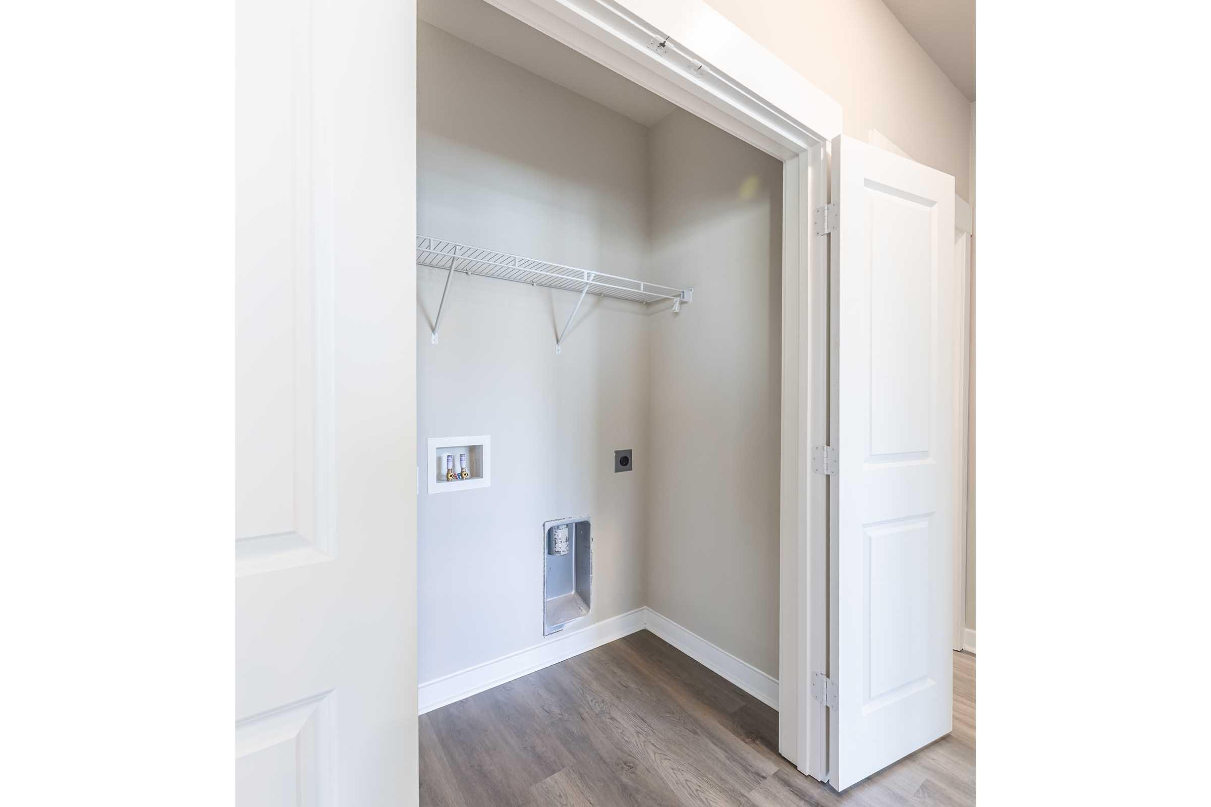 A spacious, empty closet with light-colored walls and a hardwood floor. The closet features a wire shelf along the back wall and has no doors, allowing easy access. There is an electrical outlet and a small opening on the wall, suggesting space for appliances or additional storage.