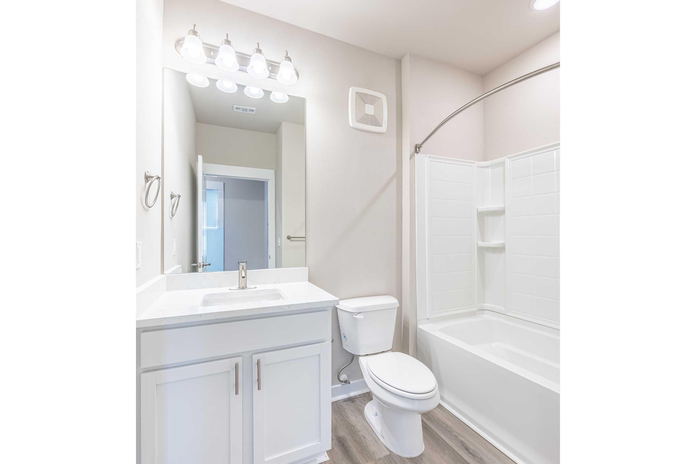 A modern bathroom featuring a white bathtub with an adjacent shower, a white toilet, and a vanity with a sink. Bright lighting is provided by four fixtures above the mirror. The walls are painted in light colors, and the flooring has a wood-like appearance.
