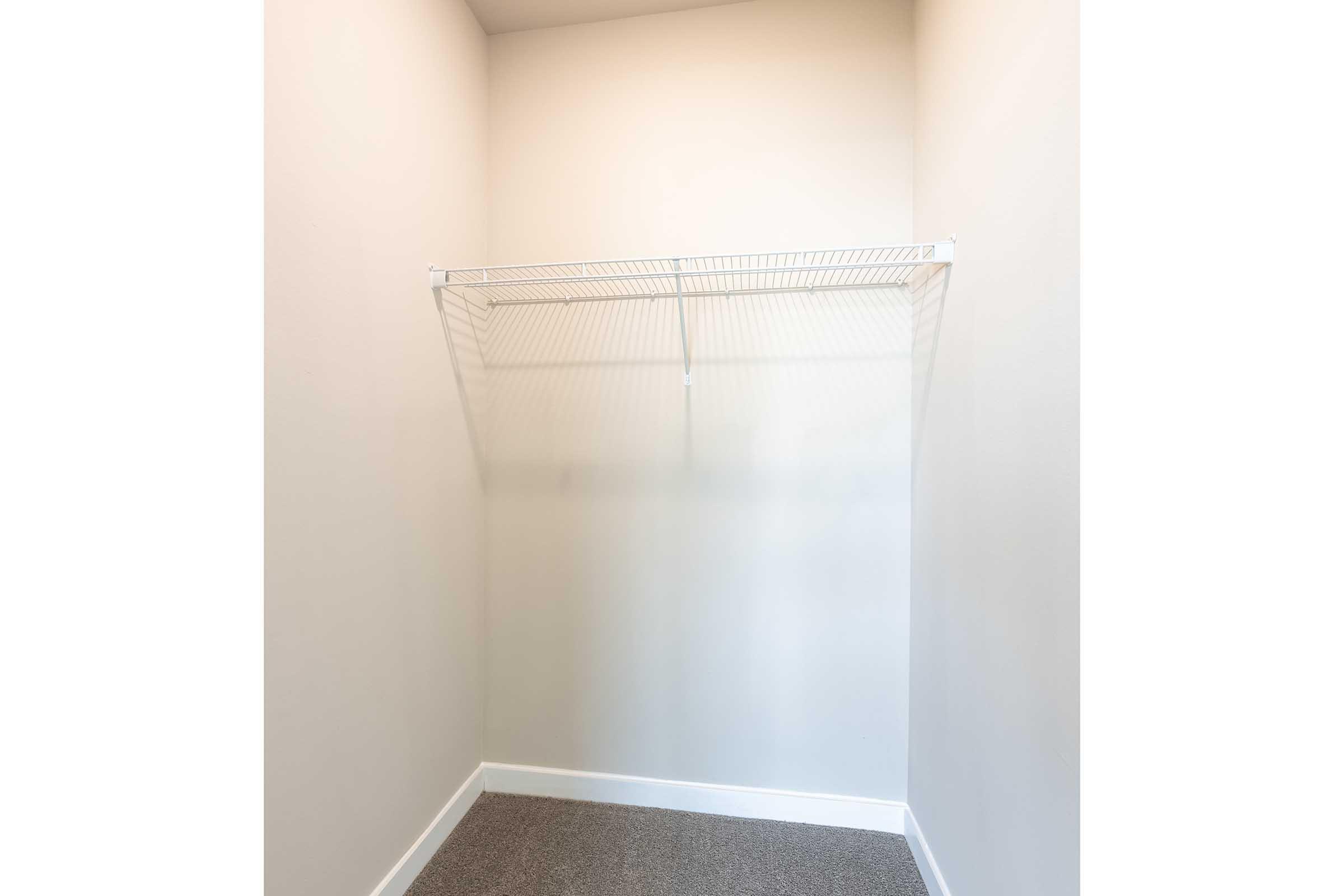 A clean, empty closet with a metal wire shelf attached to the wall. The walls and floor are neutral-colored, creating a spacious feel. The closet is well-lit with natural light, highlighting the minimalistic design.