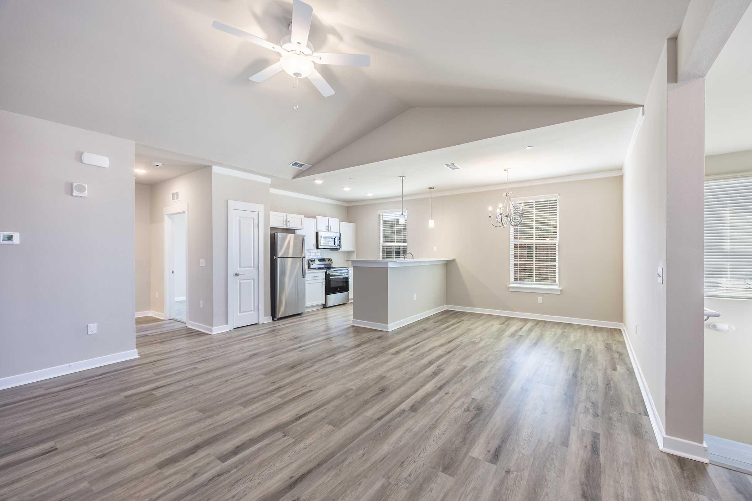 A bright and spacious living area featuring light wood flooring, a ceiling fan, and large windows. The kitchen is visible with stainless steel appliances and a bar counter. The walls are painted in a light, neutral color, creating a modern and inviting atmosphere.