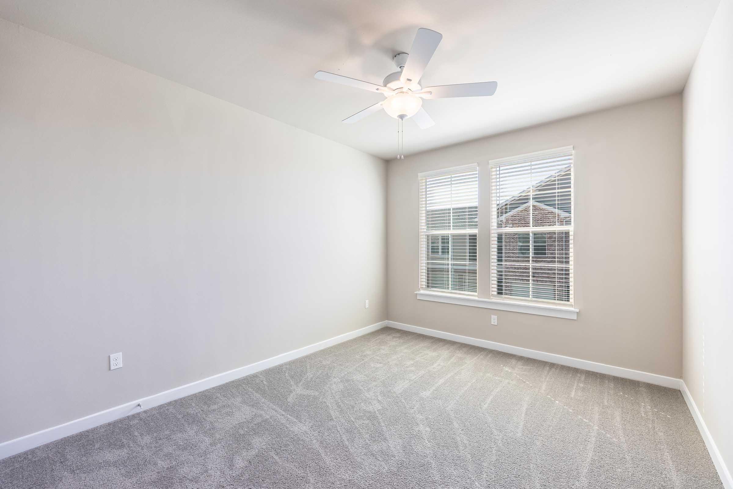 Empty room with light gray carpet, a ceiling fan, and two large windows allowing natural light. The walls are painted a soft beige, and there are no furnishings present, creating an open and airy feel.